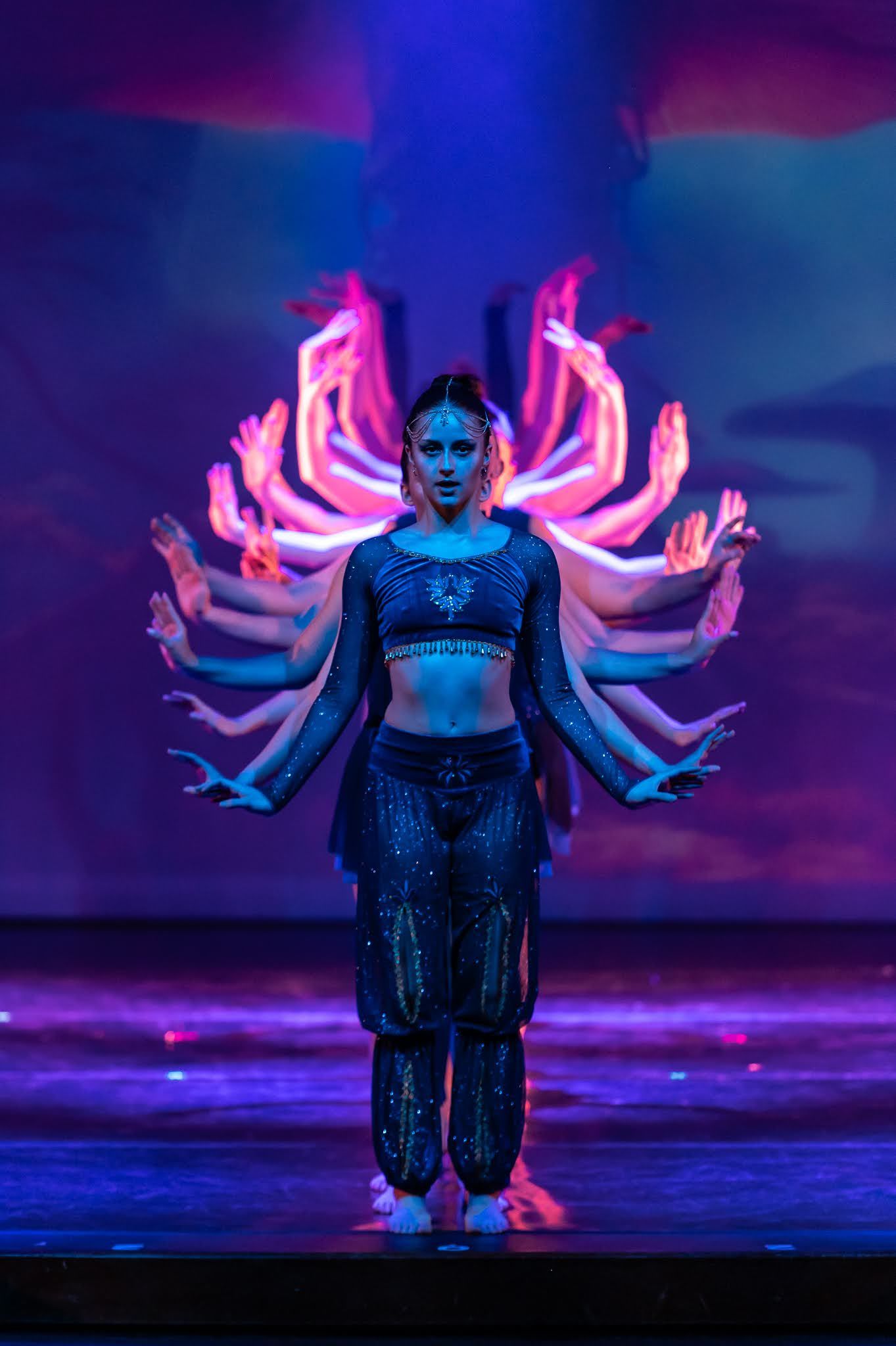 Dancer in dark costume poses center stage surrounded by multi-armed silhouette under neon lights.
