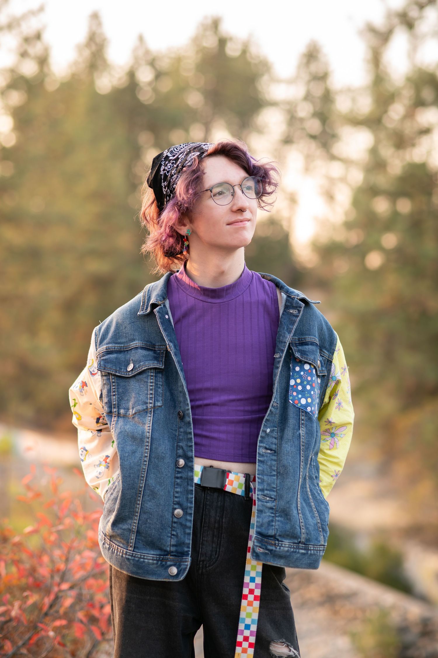 Teen with purple hair wearing denim jacket and purple shirt posing outdoors in golden hour light.
