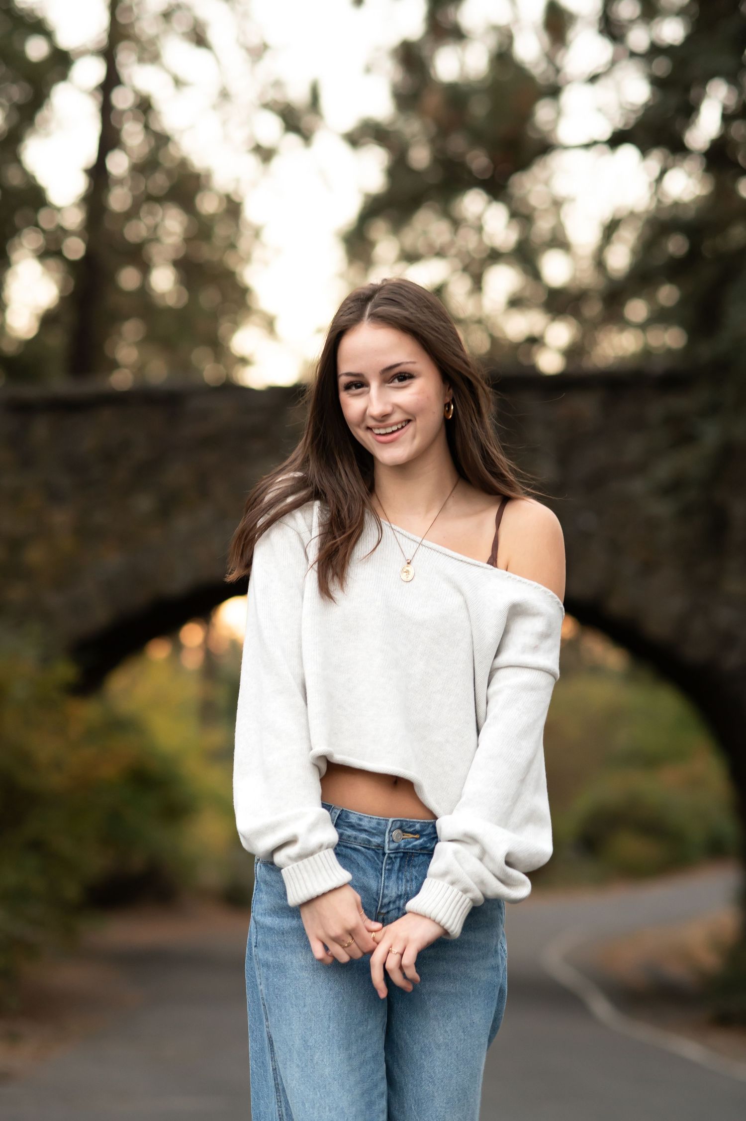 Smiling young woman in white off-shoulder crop top and jeans posing near stone bridge at sunset.