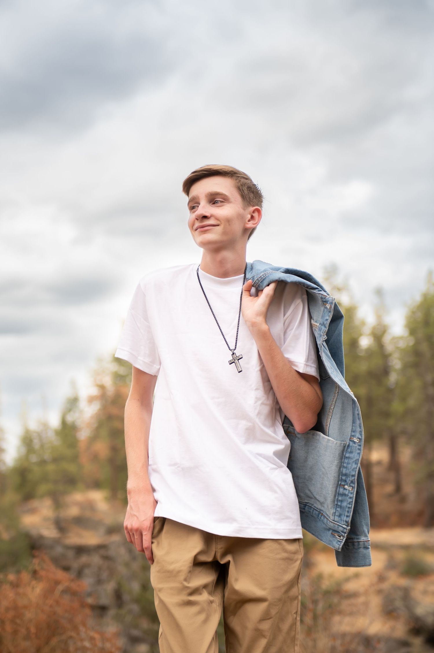 Teen boy in white t-shirt holding a denim jacket over his shoulder, wearing a cross necklace outdoors.