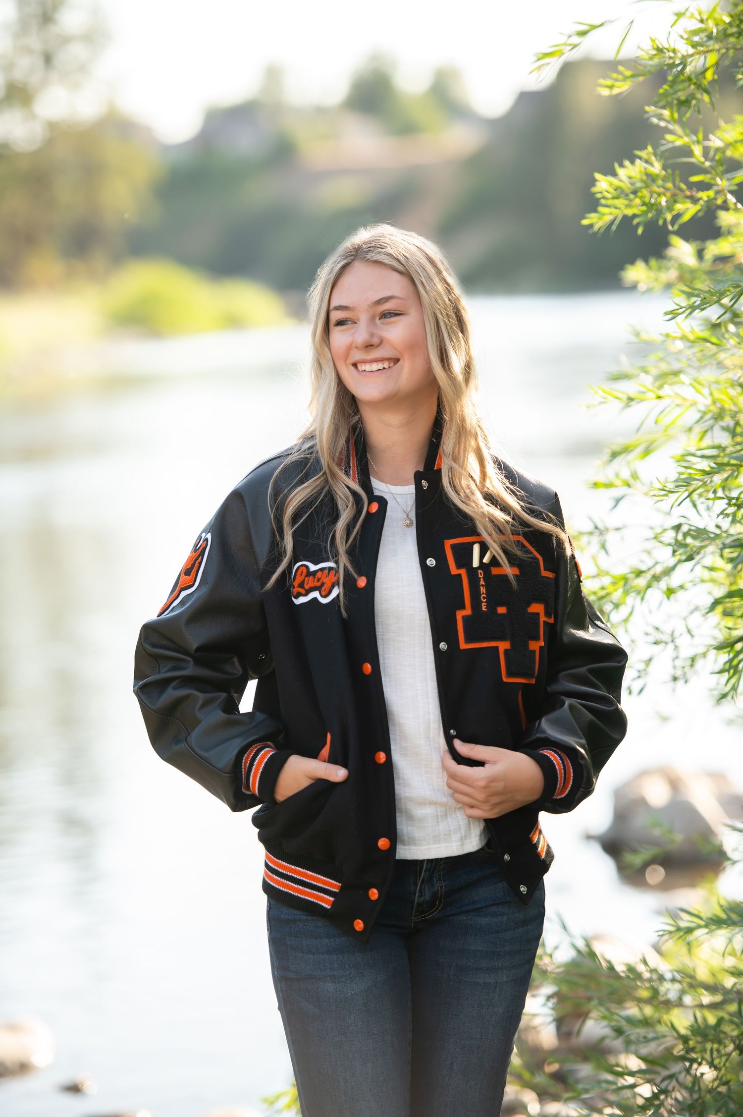Blonde teen in black varsity jacket smiles near riverside trees.