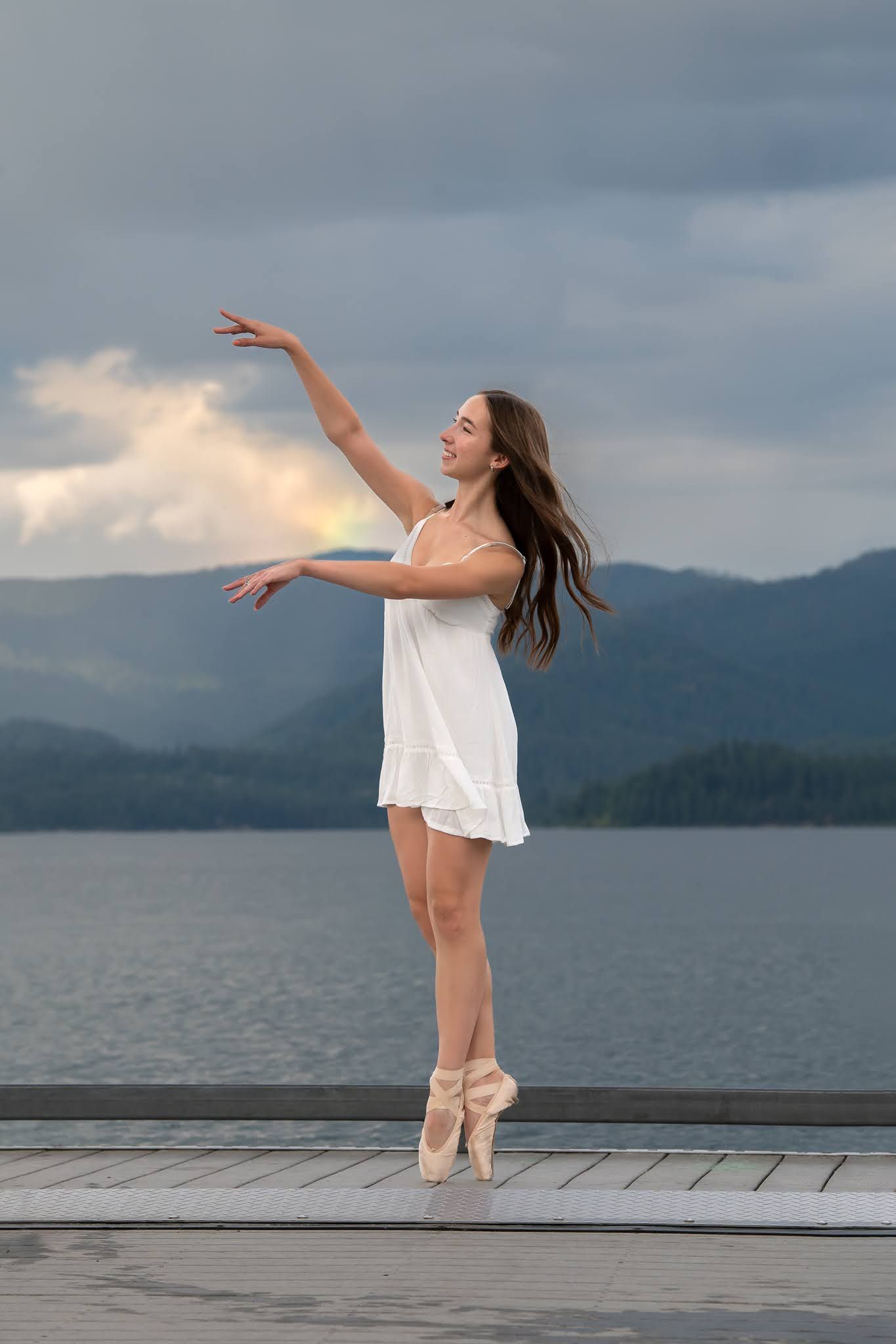 Ballerina in white dress dancing en pointe on lakeside dock with mountains and rainbow in background.