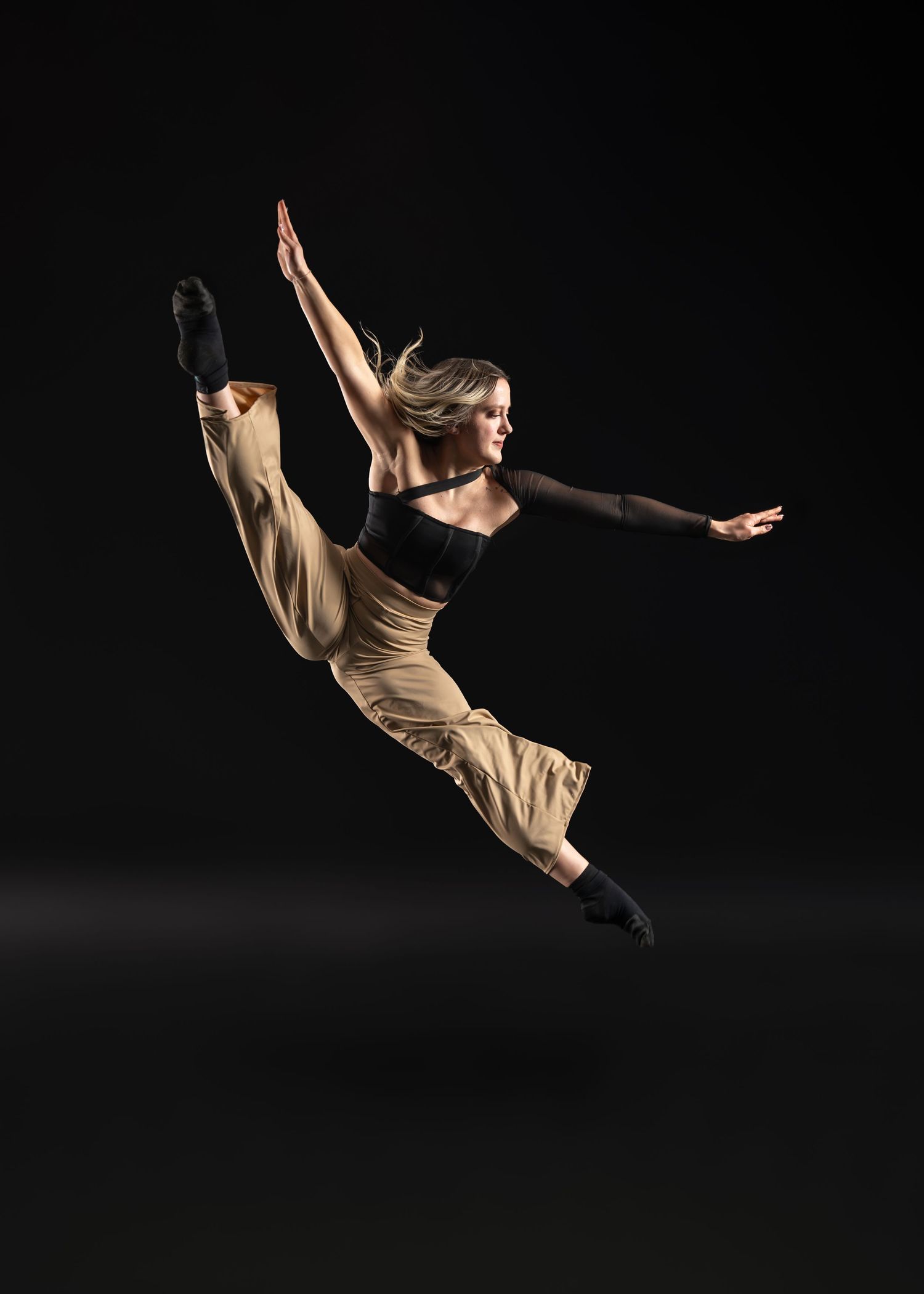 Female dancer leaping gracefully against black background in beige pants and black top, arms extended mid-air.