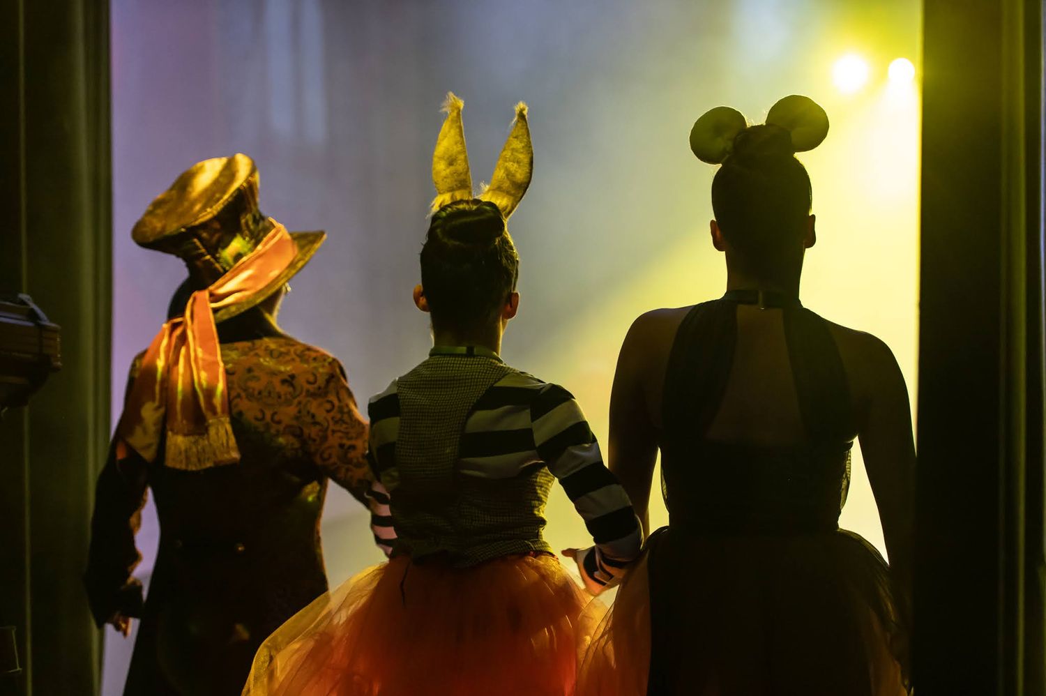 Three costumed performers silhouetted in doorway wearing top hat, bunny, and mouse ears, representing characters from Alice in Wonderland.