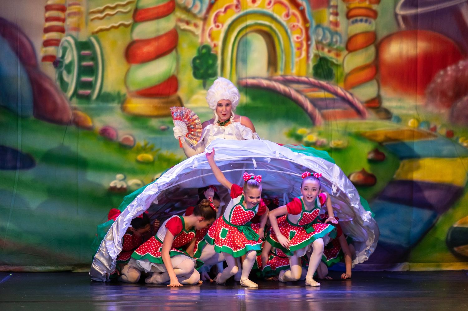 Stage performance from the ballet, The Nutcracker, with young dancers in red costumes emerging from under large white hoop skirt dress.