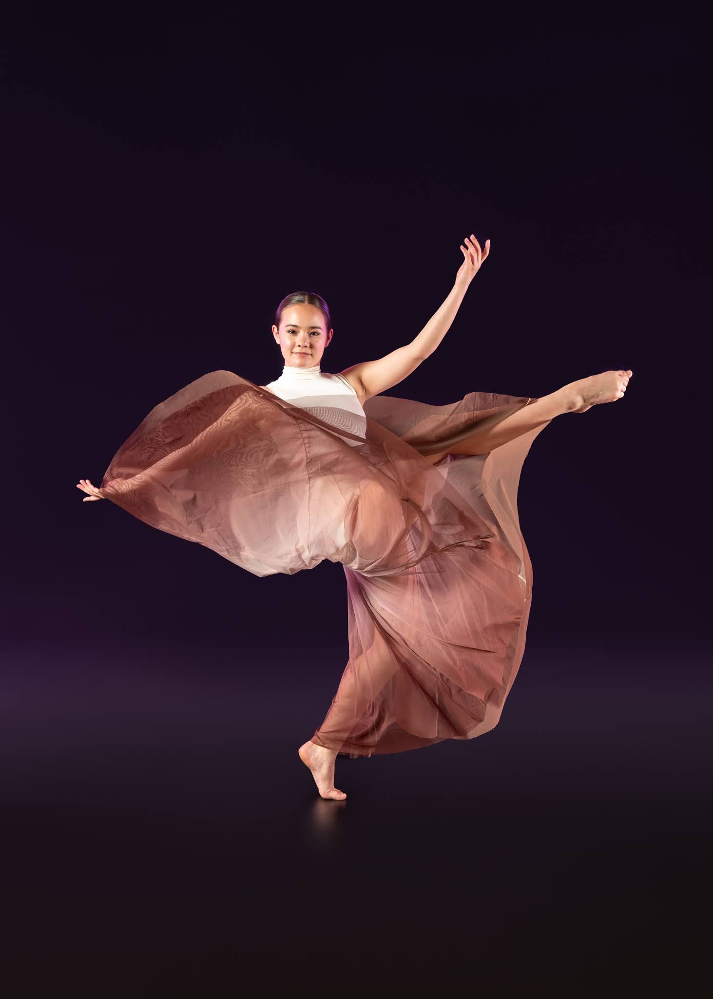 Dancer in flowing brown skirt performs graceful contemporary routine on dark stage.