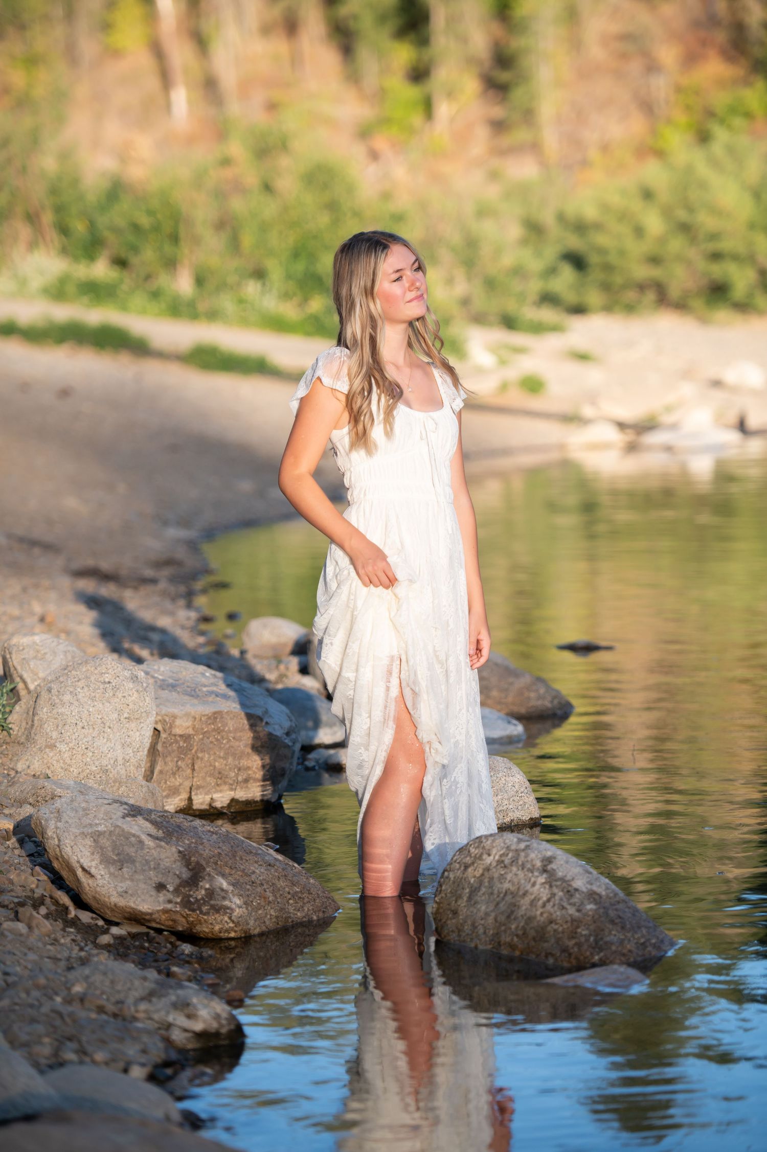 Young blonde woman in white lace dress stands in shallow river.