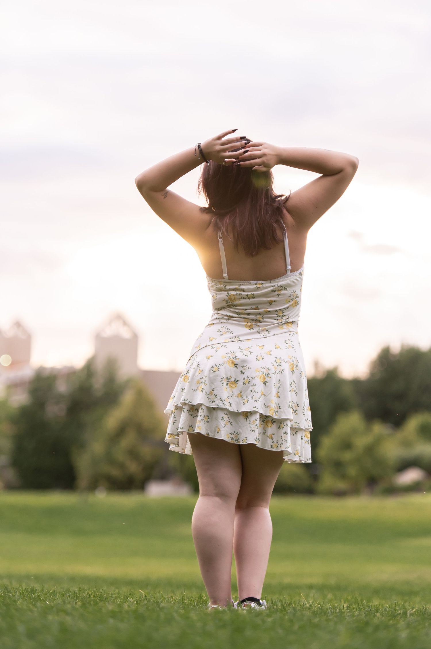 Young woman in floral sundress standing in green park field with hands in hair at sunset.