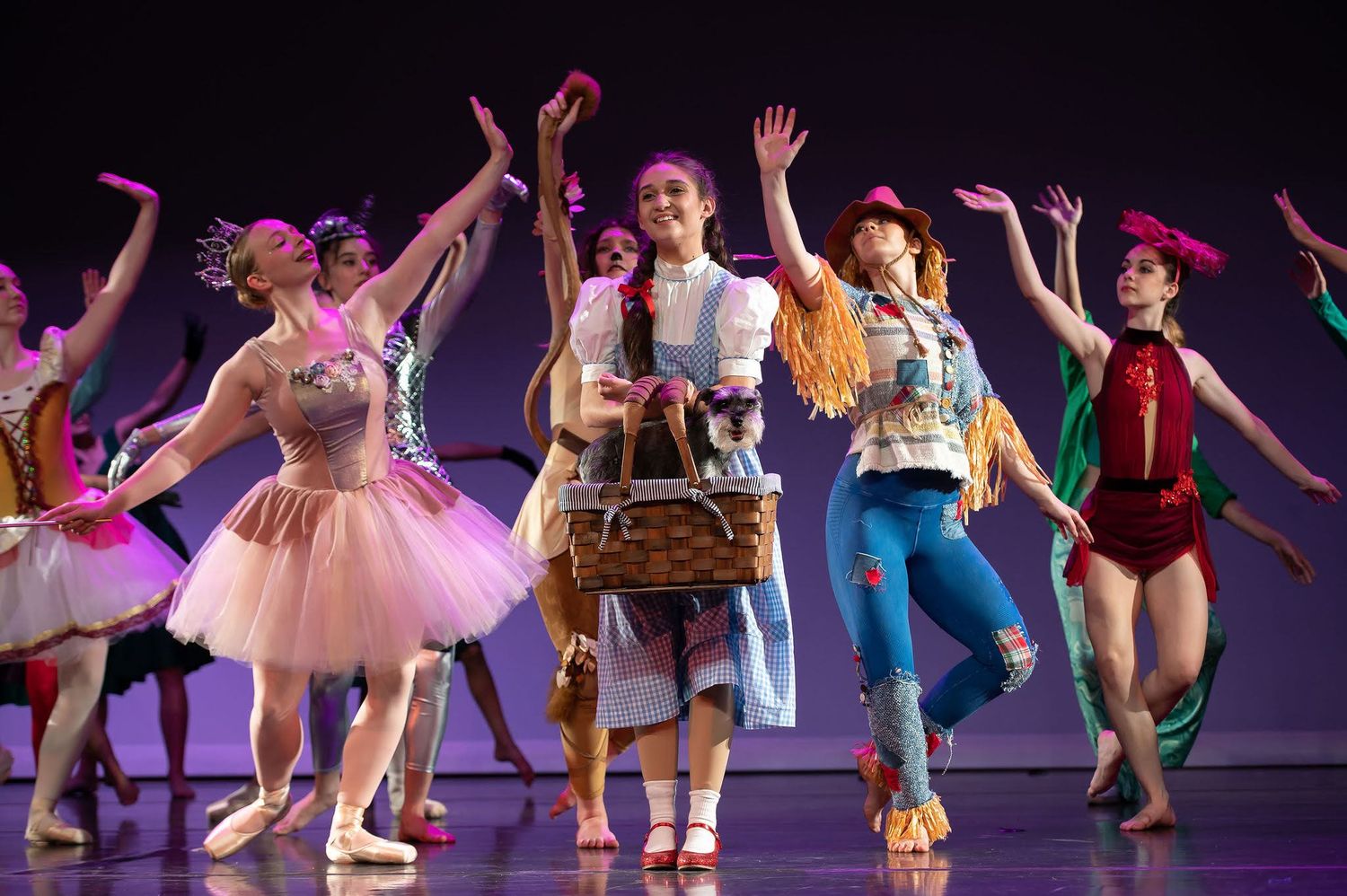 Dancers in colorful costumes wave goodbye to Dorothy in a lively Wizard of Oz themed ballet recital on stage.