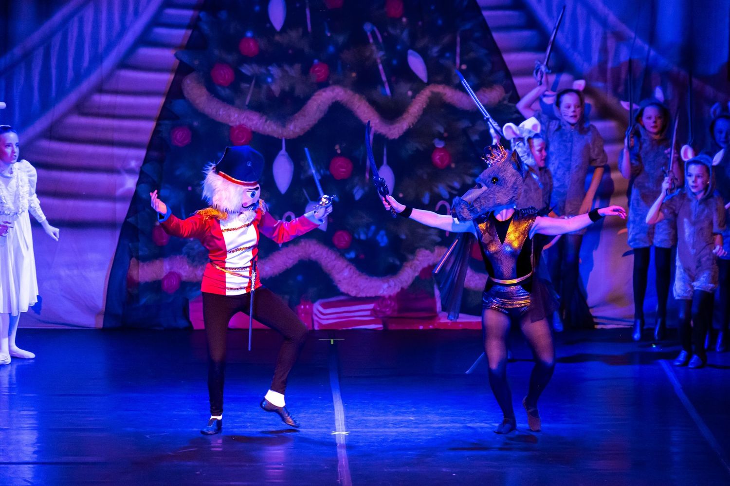 Two performers in Nutcracker costumes duel on a festive stage with Christmas tree and ensemble cast backdrop.