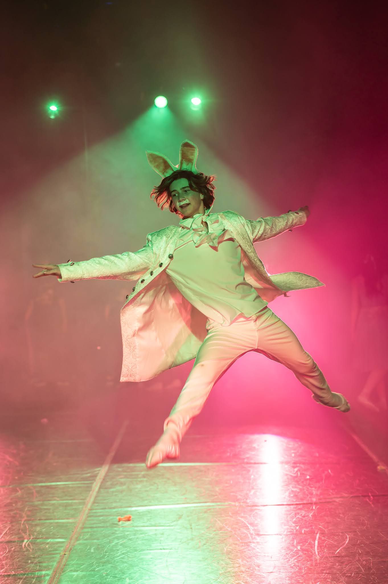 Performer in white costume and bunny ears leaps dramatically on stage under colorful green and pink stage lights.