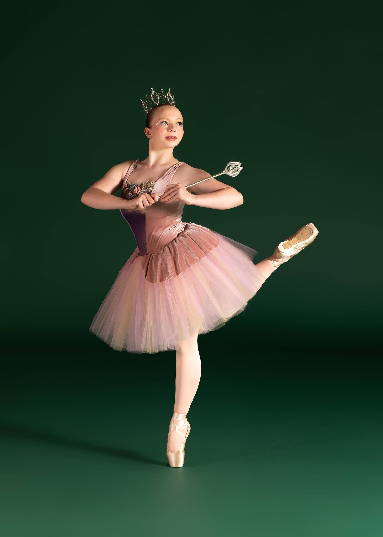 Ballerina in pink tutu and crown poses on pointe holding a wand on green background.