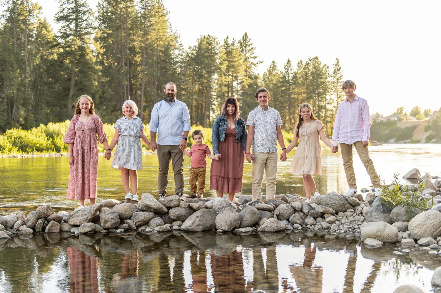 Large family group portrait standing on rocky riverbank surrounded by tall pine trees at golden hour.