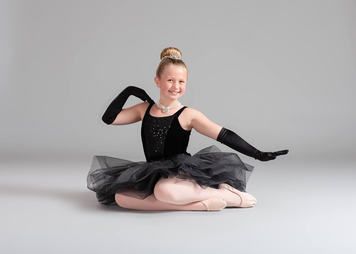 Smiling young girl in black ballet tutu and gloves sits in a split pose on a gray studio background.