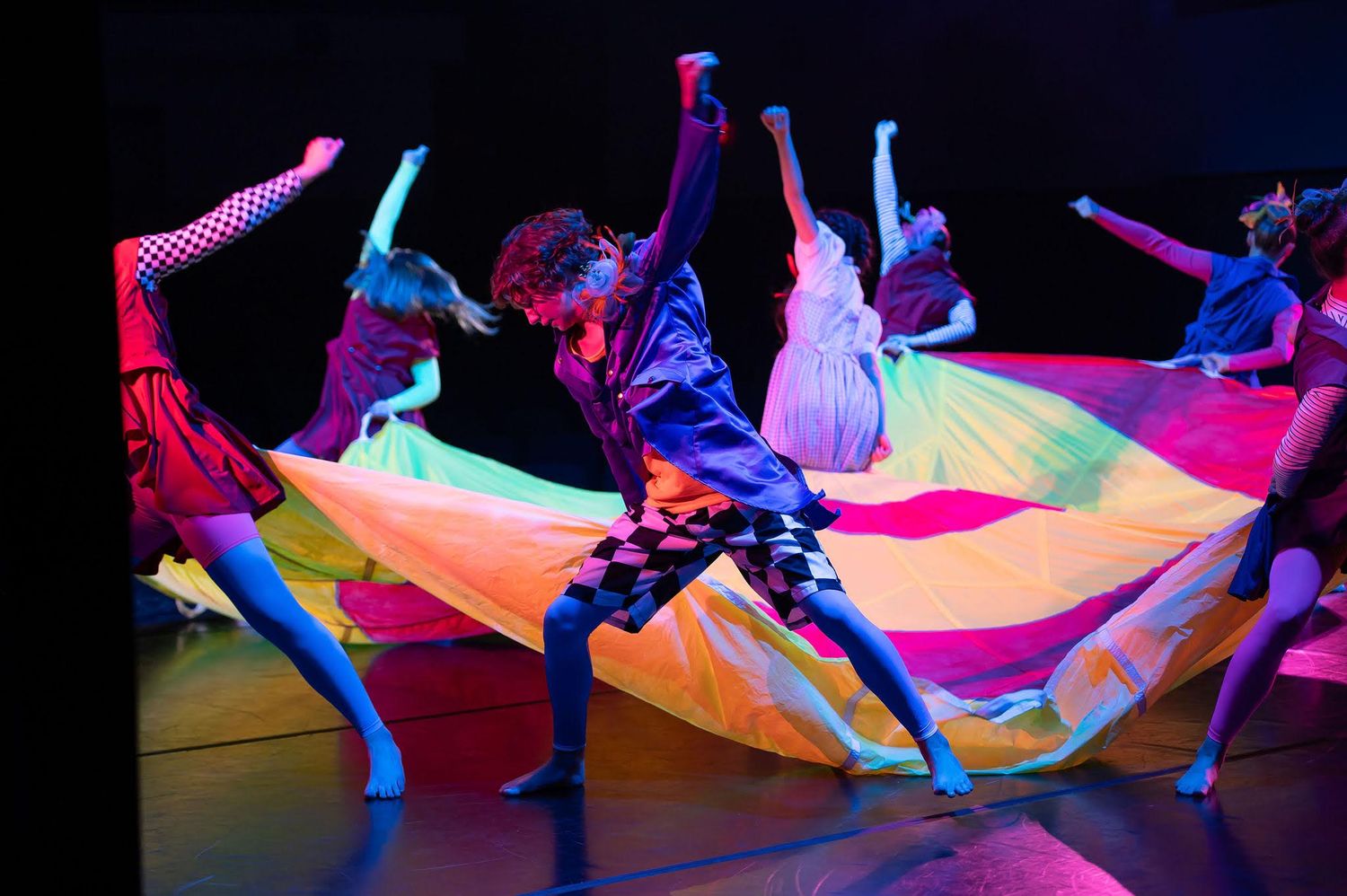 Young dancers perform with a colorful rainbow parachute under vibrant stage lighting.