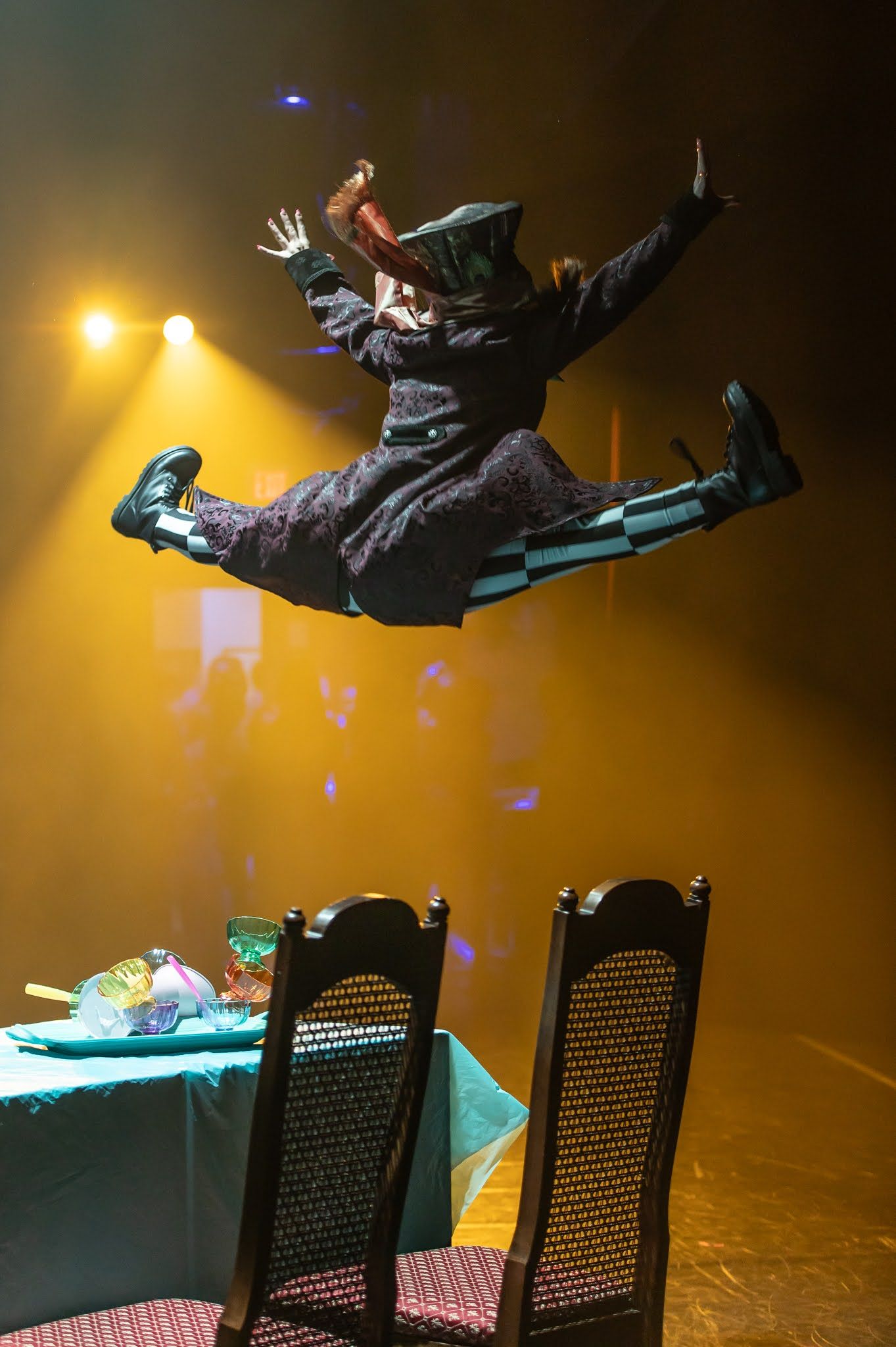 Performer in Mad Hatter costume leaps dramatically above a tea party table on stage.