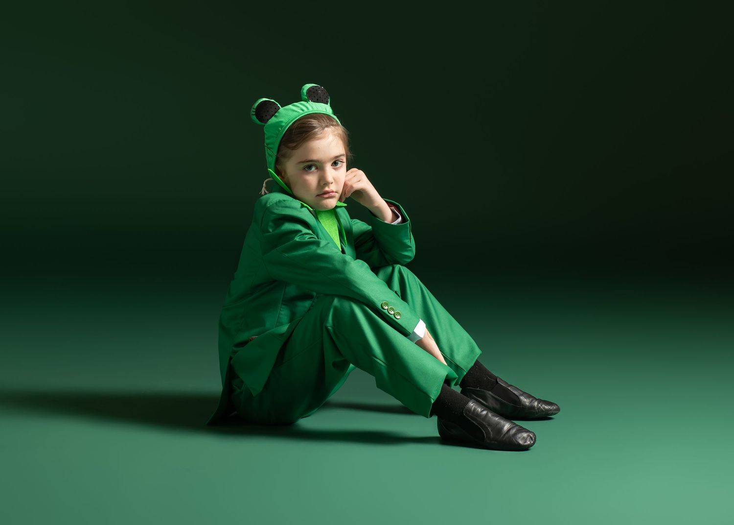 Child performer sits on green floor wearing full green frog costume with hood and black boots.