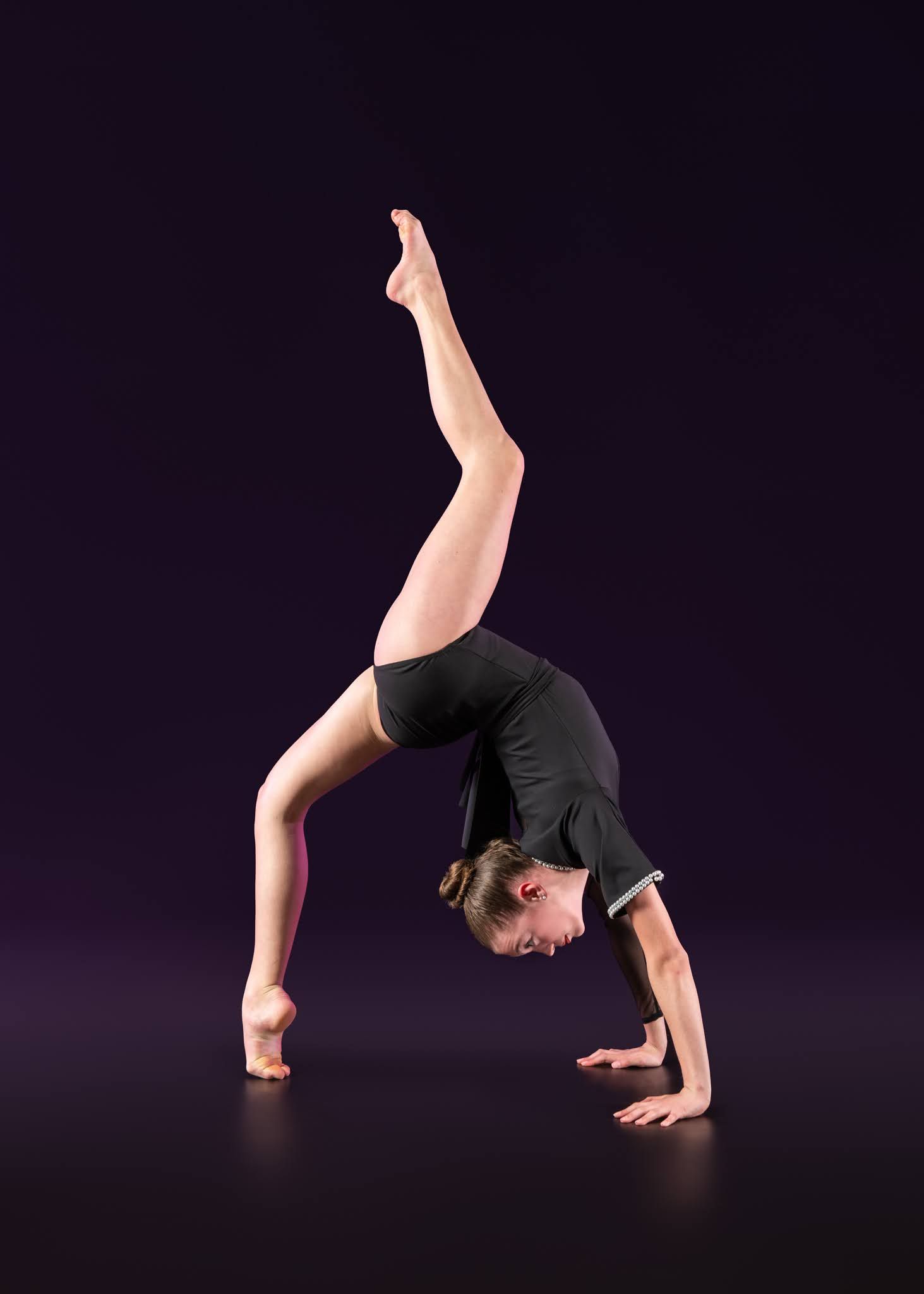 Gymnast in black leotard executes a one-handed cartwheel with leg extended on dark background.
