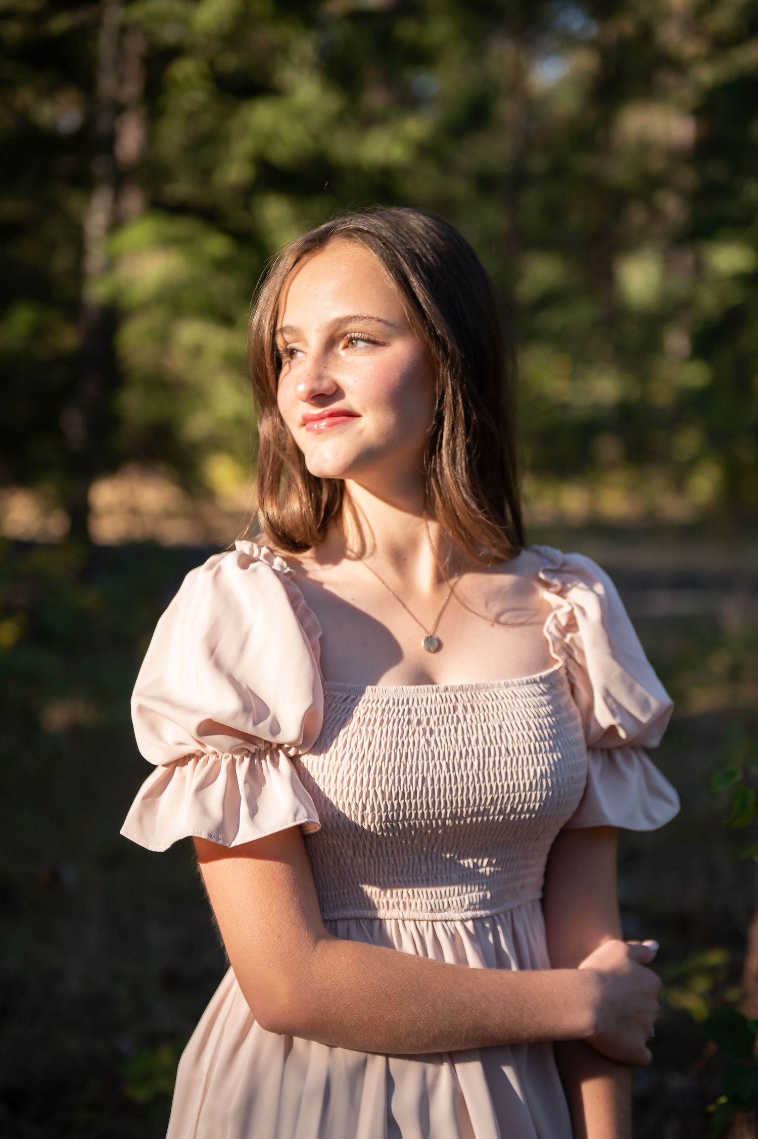 Young woman in cream smocked dress gazing into distance with sunlit pine trees in the background.