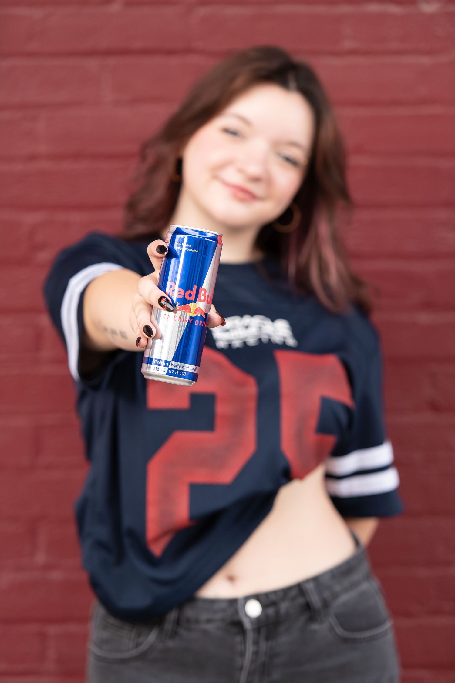 Young woman in sports jersey holding Red Bull energy drink toward camera against brick wall.