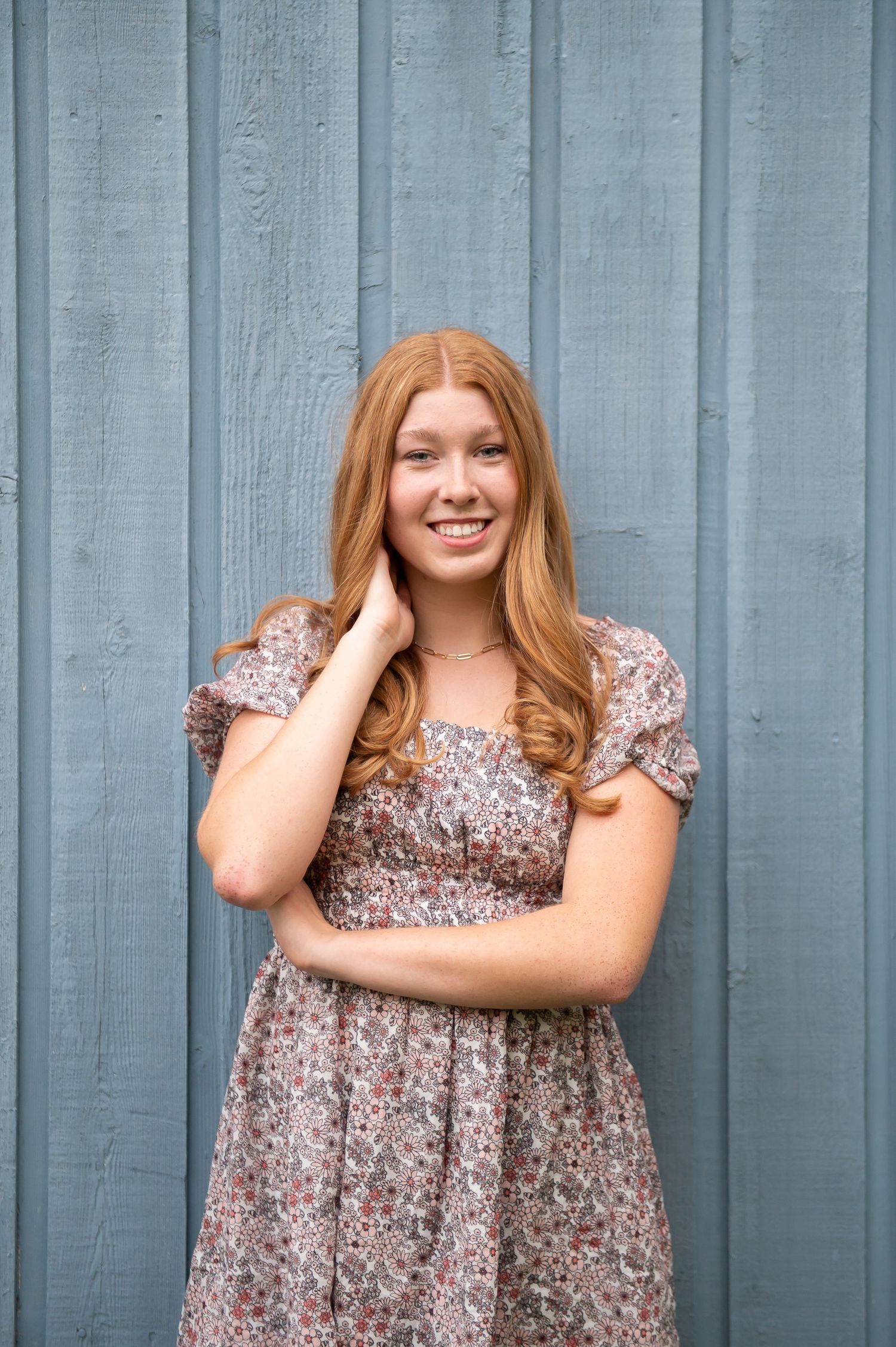 Smiling young woman with long red hair in a floral dress posing against a blue wooden wall.
