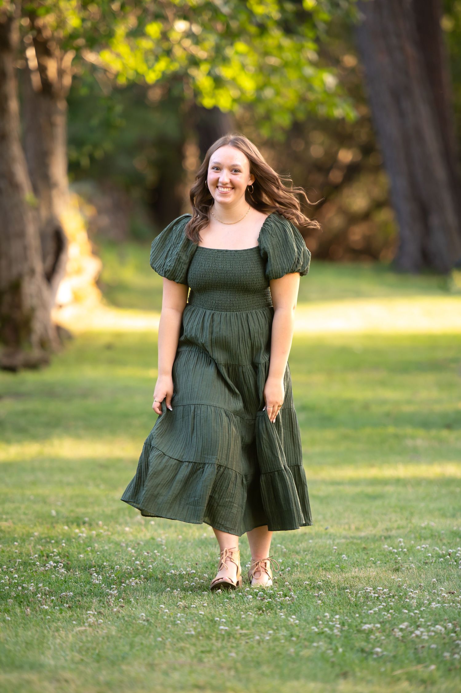 Young woman in dark green midi dress walks through sunlit park smiling.