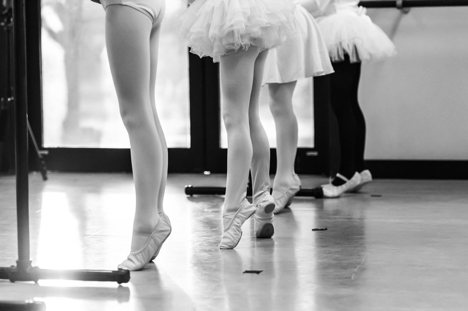 Close-up black and white photo of young ballet students feet in ballet shoes at a barre during class.