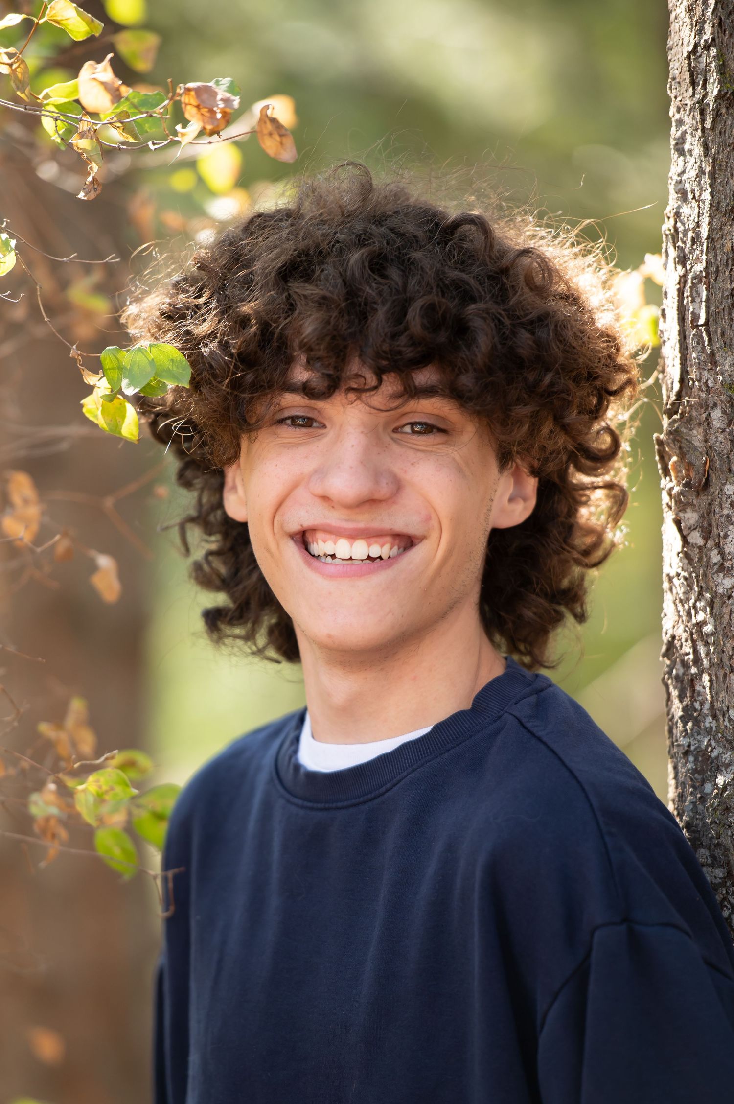 Smiling teenage boy with curly brown hair wearing a navy sweatshirt, posing outdoors near a tree with autumn leaves.