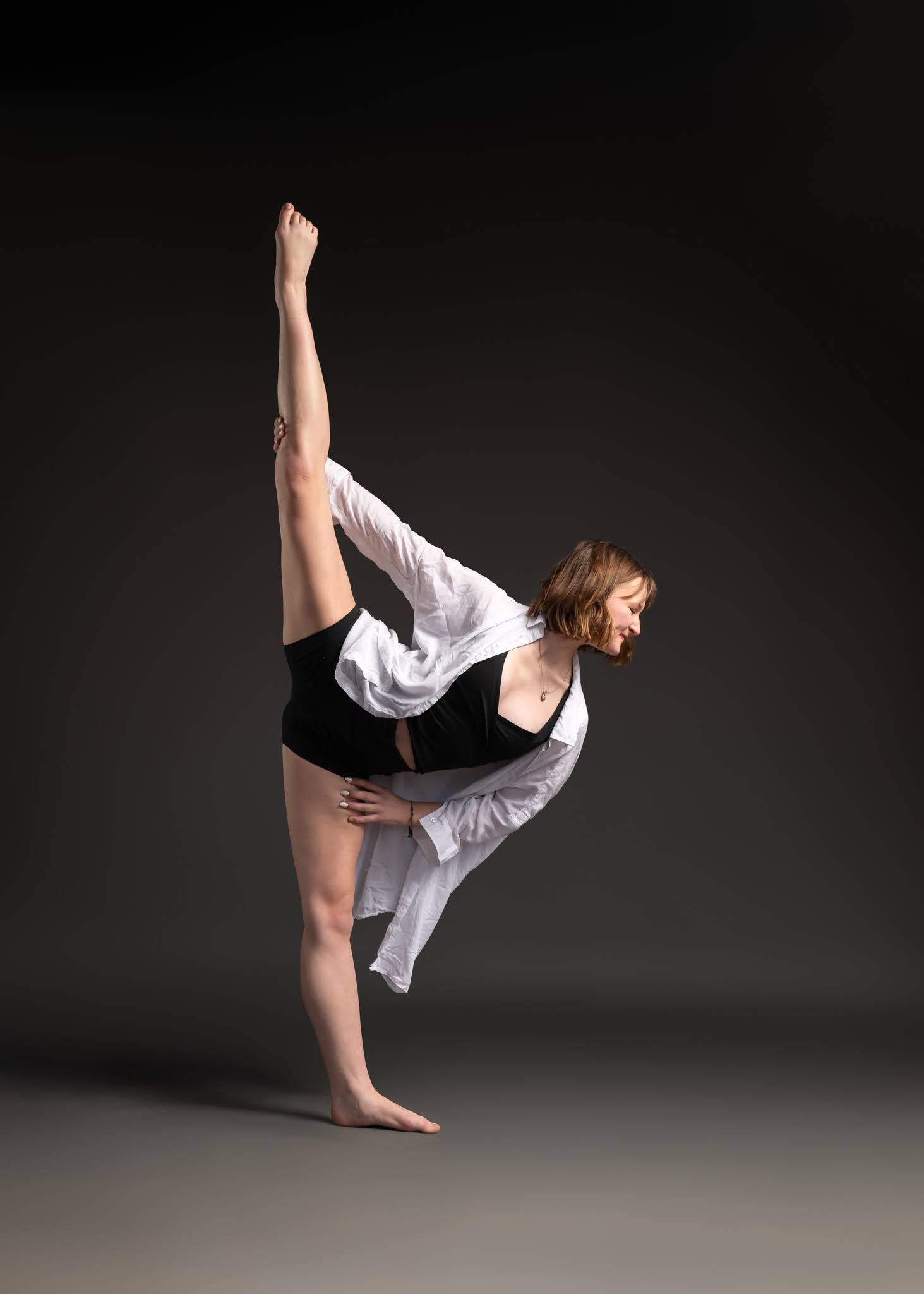 A contemporary dancer in black shorts and white shirt performs a standing split against a dark gray background.