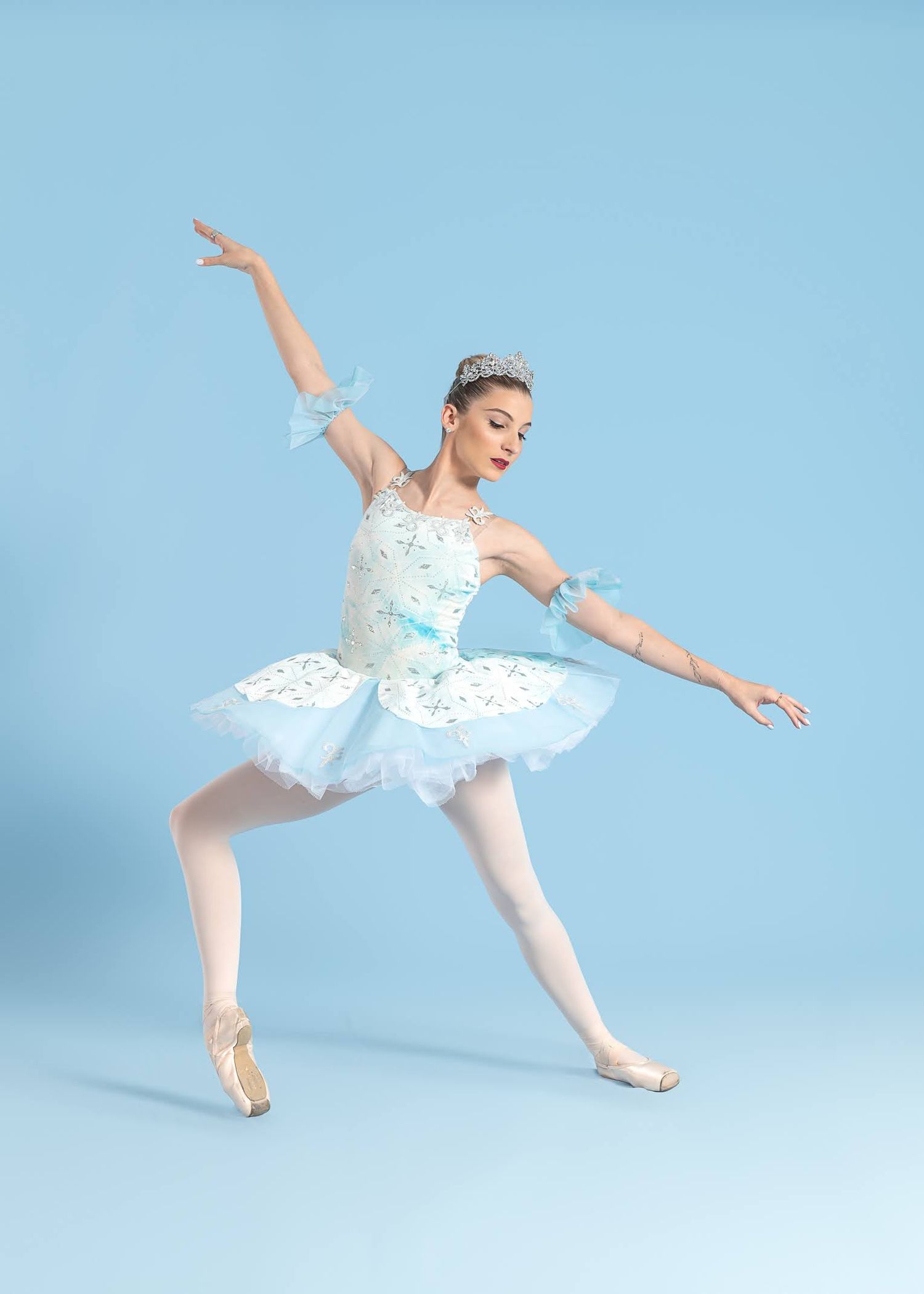 A ballerina in a white and blue tutu with tiara performs an arabesque en pointe against a light blue background.