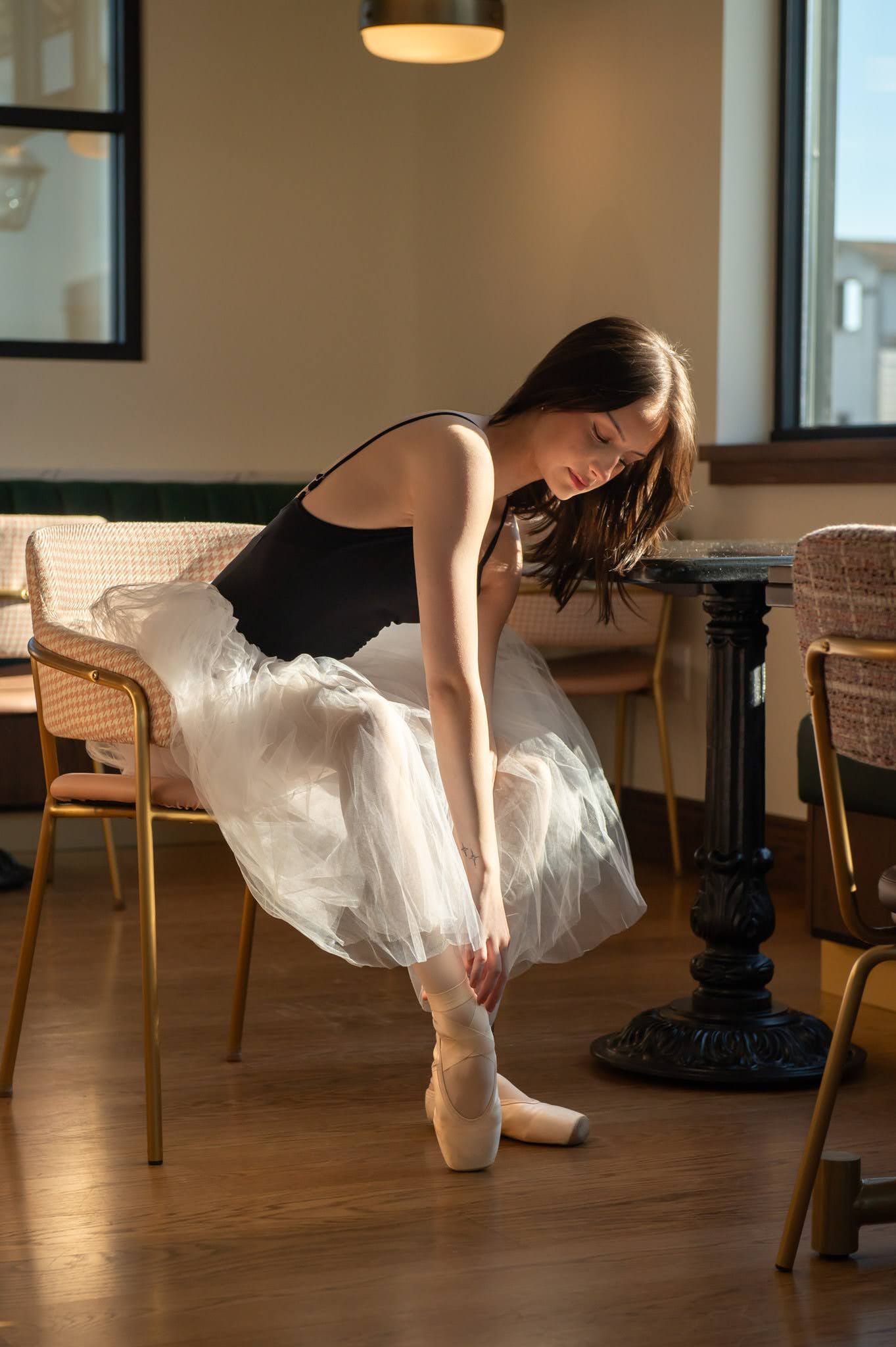 Young ballerina in black leotard and white tutu adjusting pointe shoes in sunlit cafe interior.
