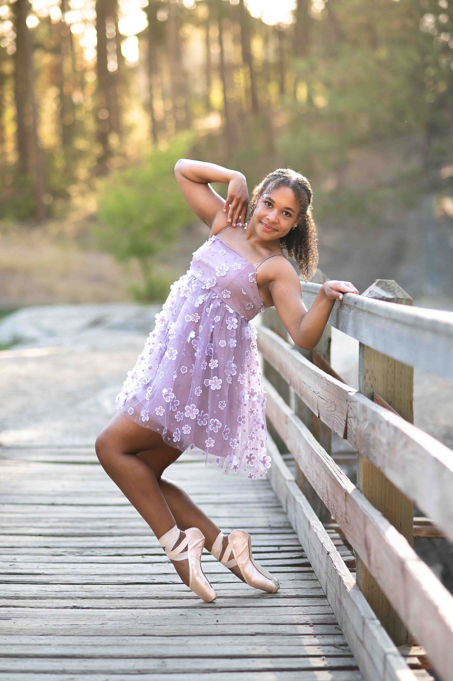 Young ballerina in lavender floral dress posing on pointe shoes on a wooden bridge in a forest.