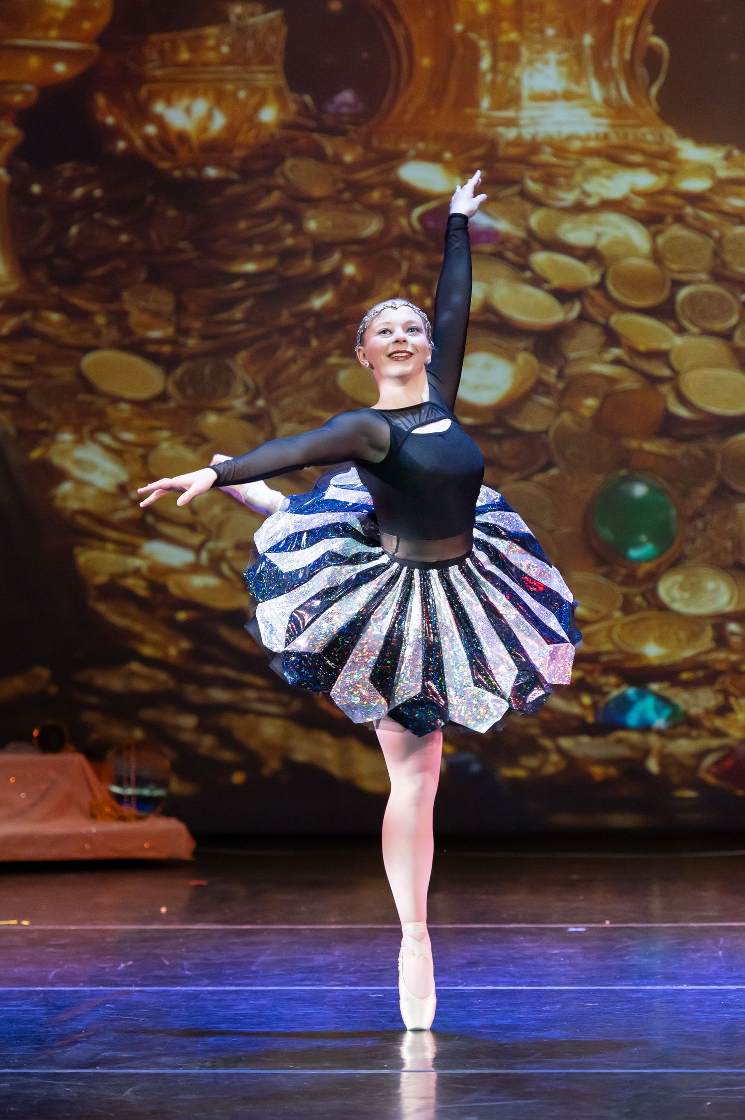 Ballerina in black and silver sequined tutu performs an arabesque on pointe before a golden treasure backdrop.