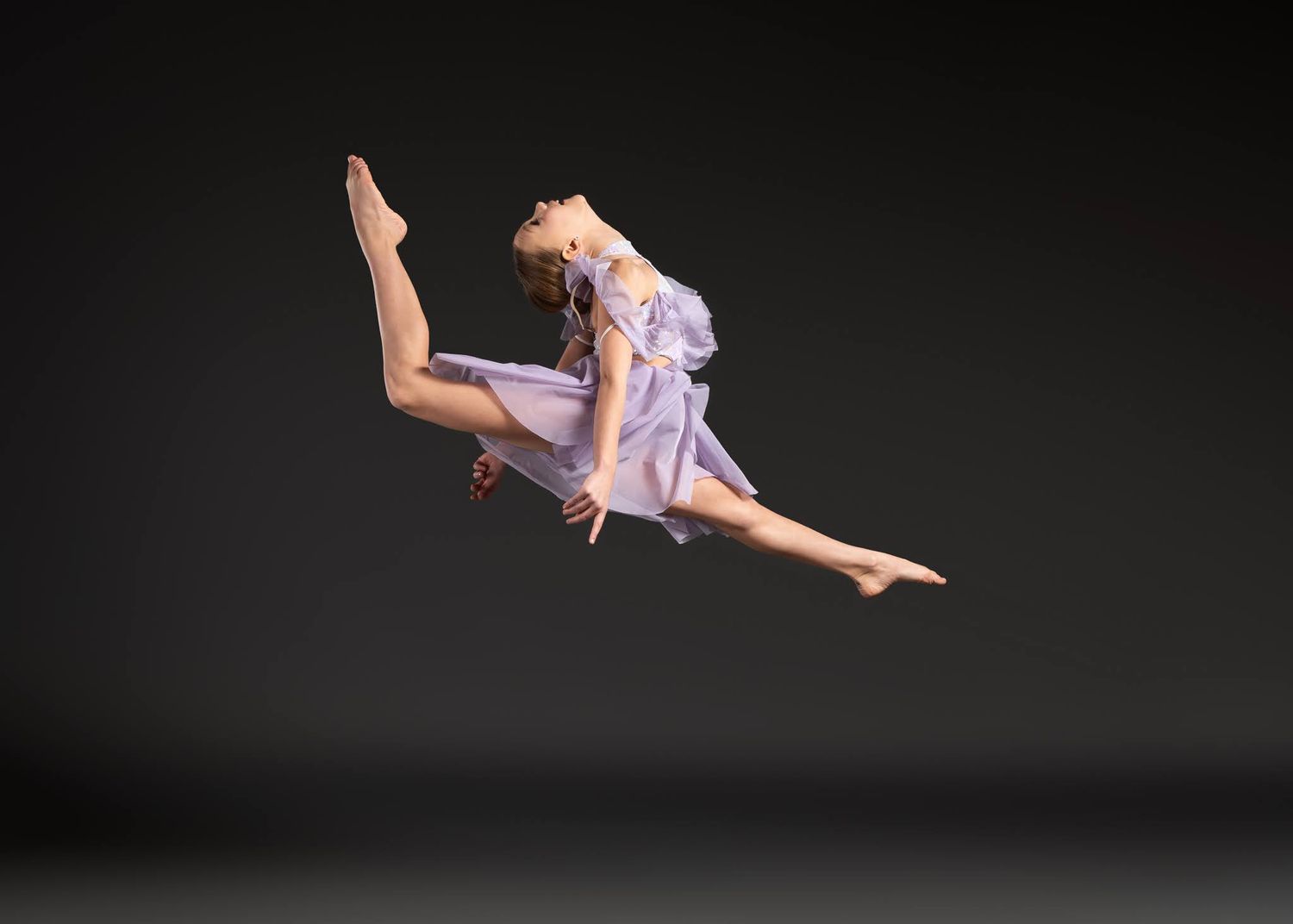 Young dancer in purple dress executing a perfect split leap against gray background.