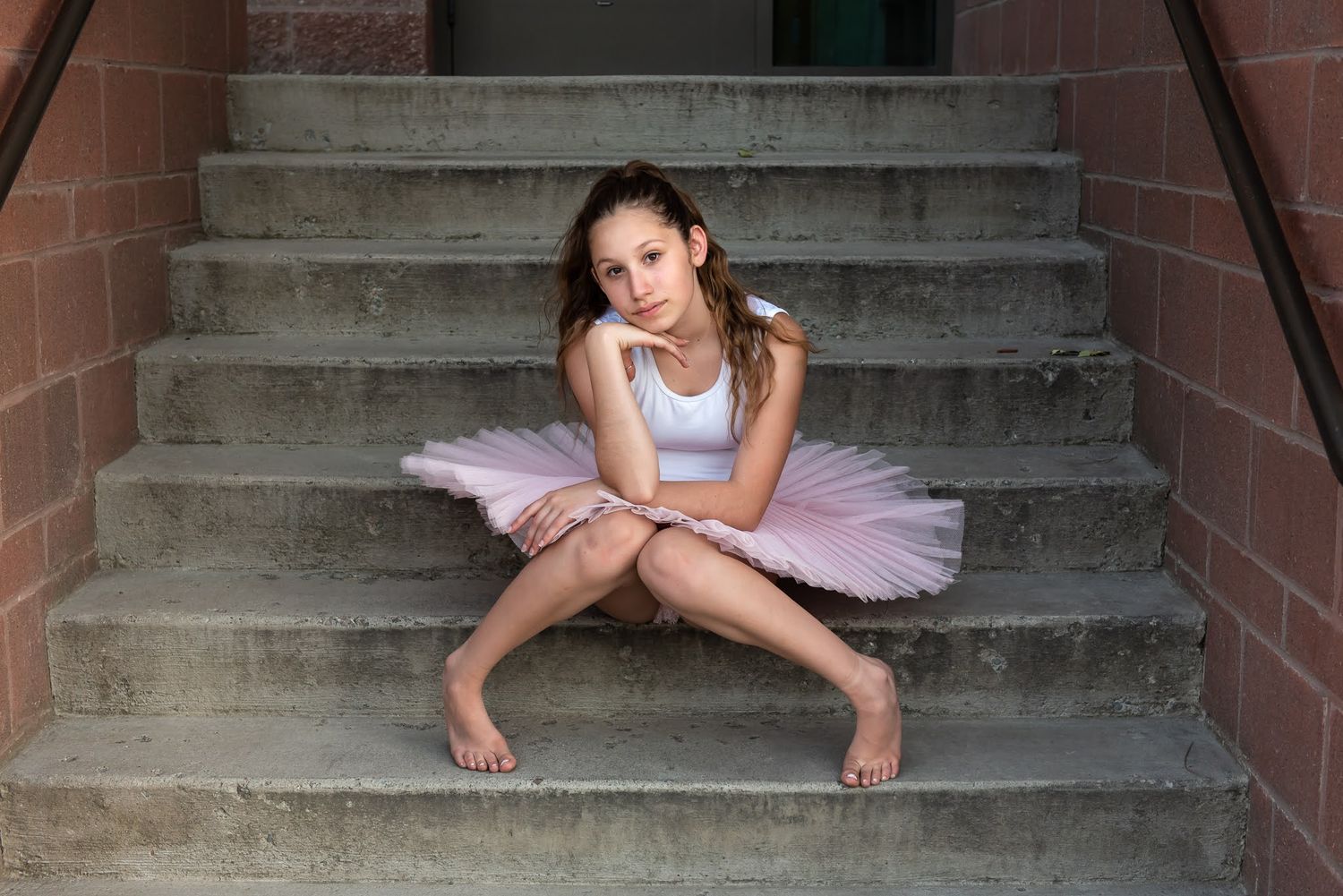 Young ballerina in pink tutu sitting on concrete stairs with a contemplative expression, barefoot.