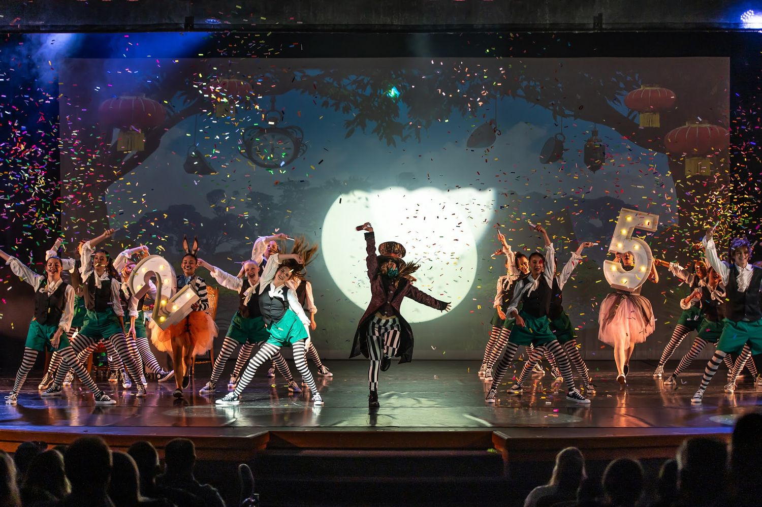 Large ensemble of dancers performs colorful stage production with vibrant backdrop and confetti.