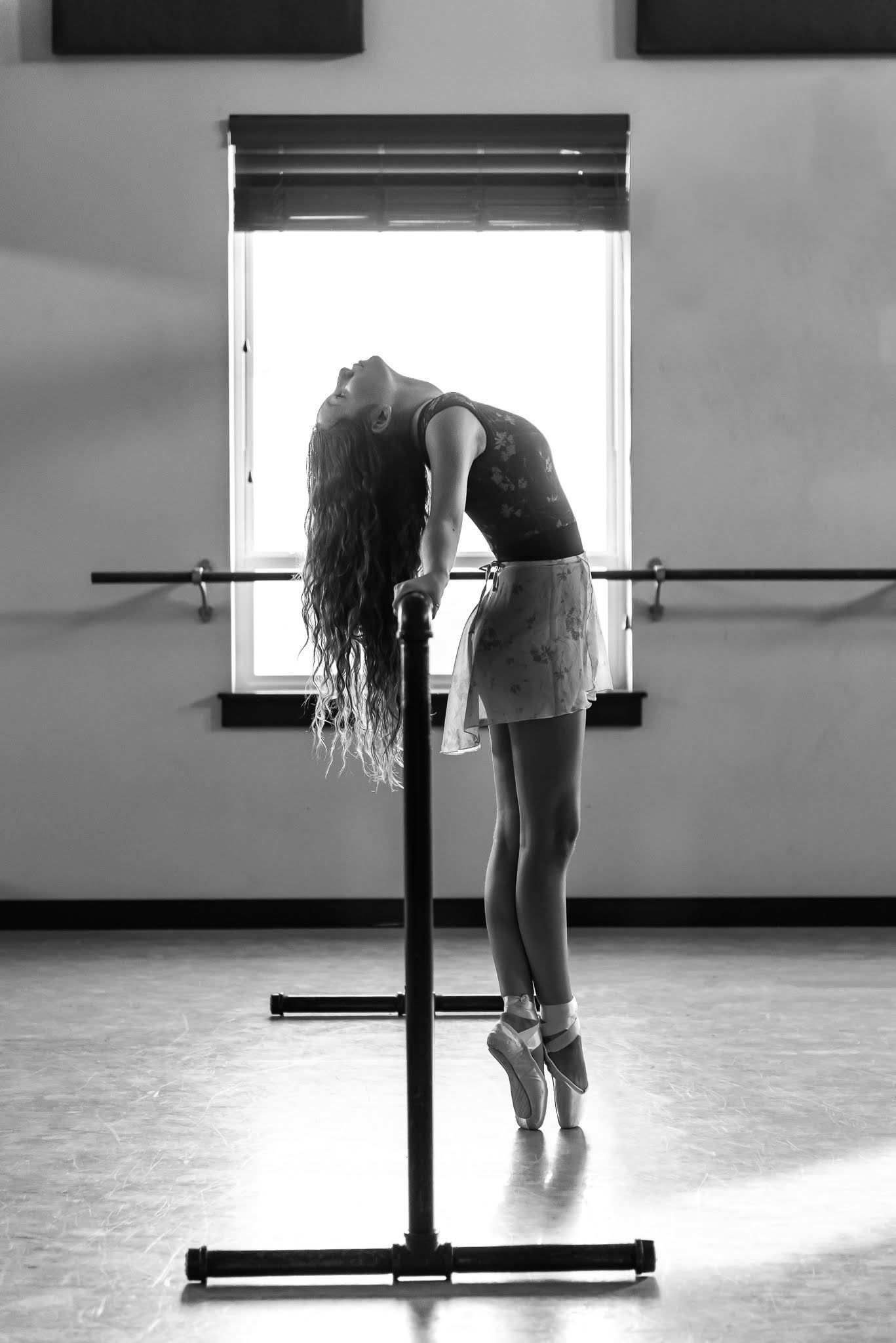 Black and white photo of ballerina en pointe at barre bending back gracefully in sunlit dance studio.