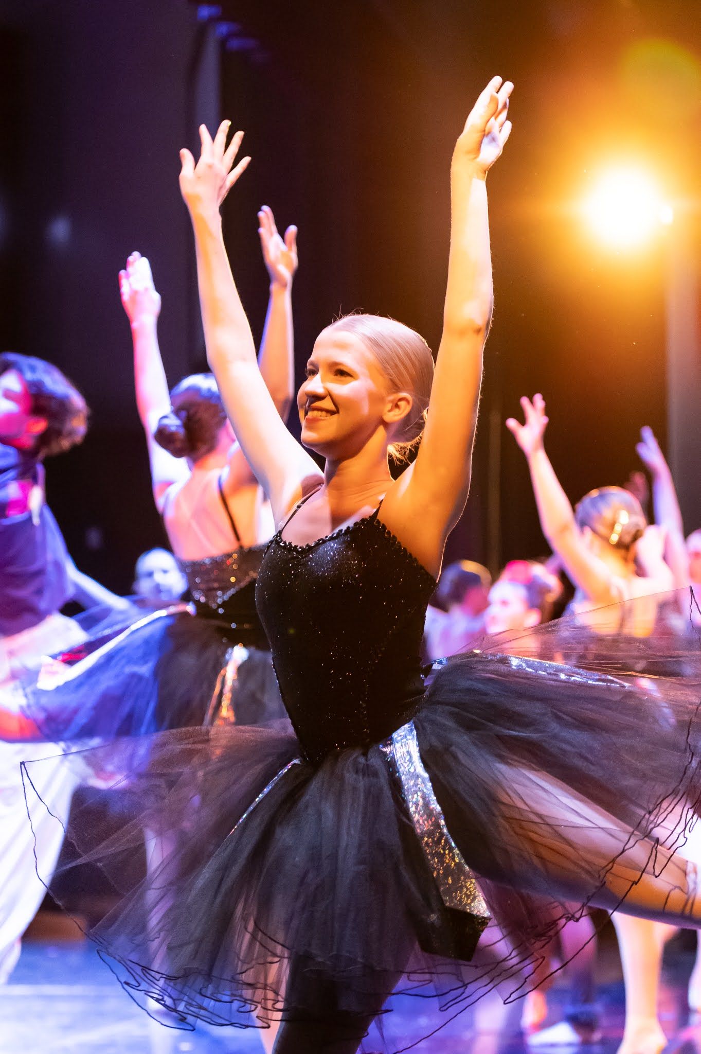 A smiling female ballet dancer in a black tutu raises her arms triumphantly under warm stage lighting during a performance.