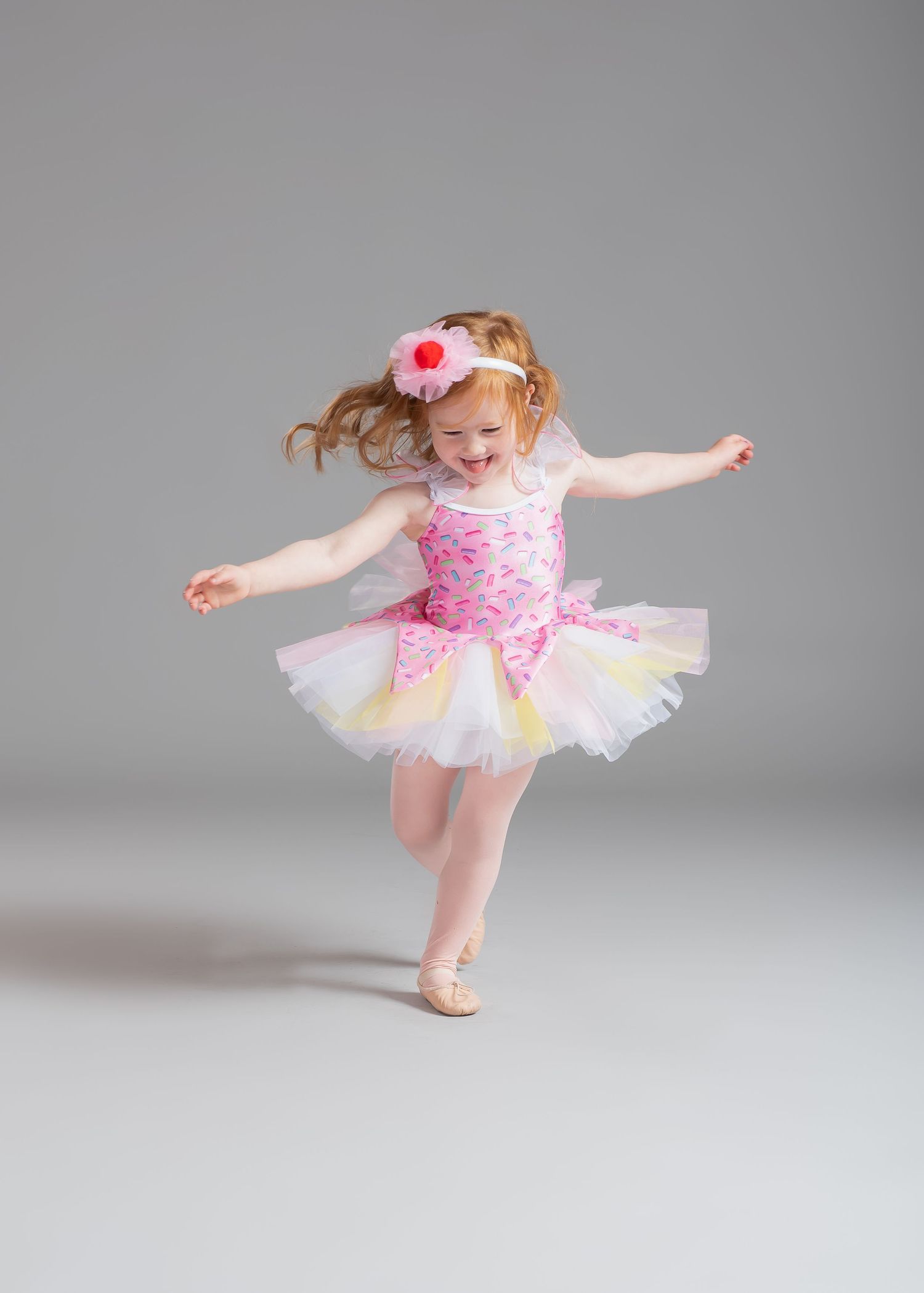 Joyful young girl in pink tutu ballet costume dances freely on gray background.
