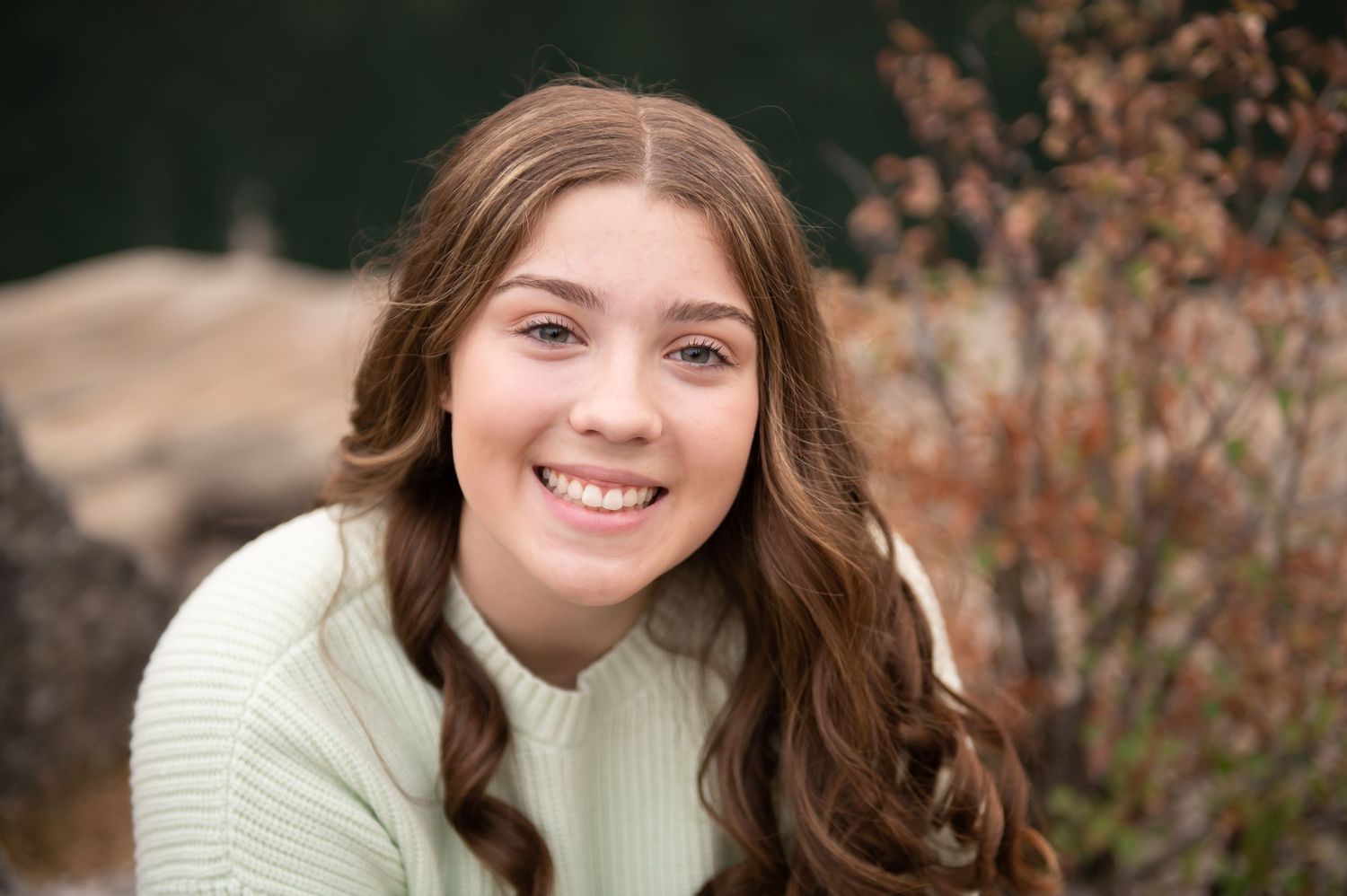 Smiling teenage girl with long wavy brown hair wearing a mint green sweater in an outdoor setting.