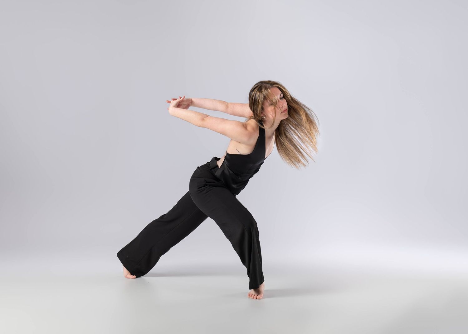Contemporary dancer in black outfit performs dynamic lunge with flowing hair on light gray background.
