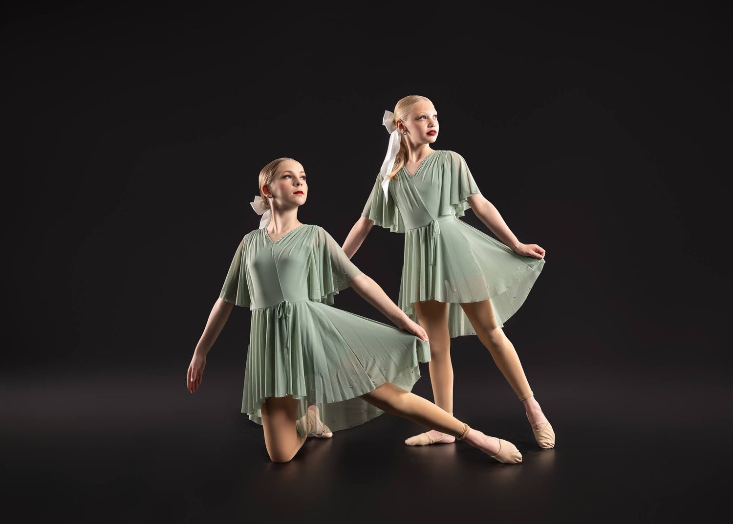 Two dancers in sage green dresses perform an elegant duet on dark studio background.