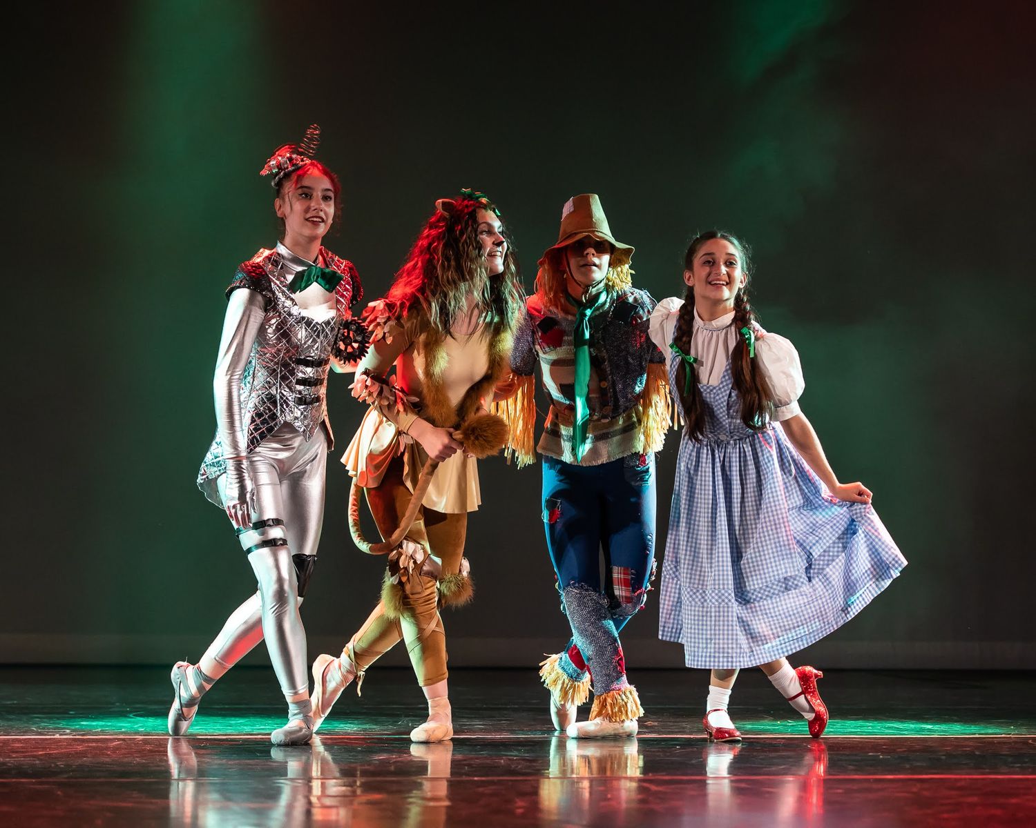 Four dancers in Wizard of Oz costumes including Dorothy, Tin Man, Cowardly Lion, and the Scarecrow perform on a theatrical stage.