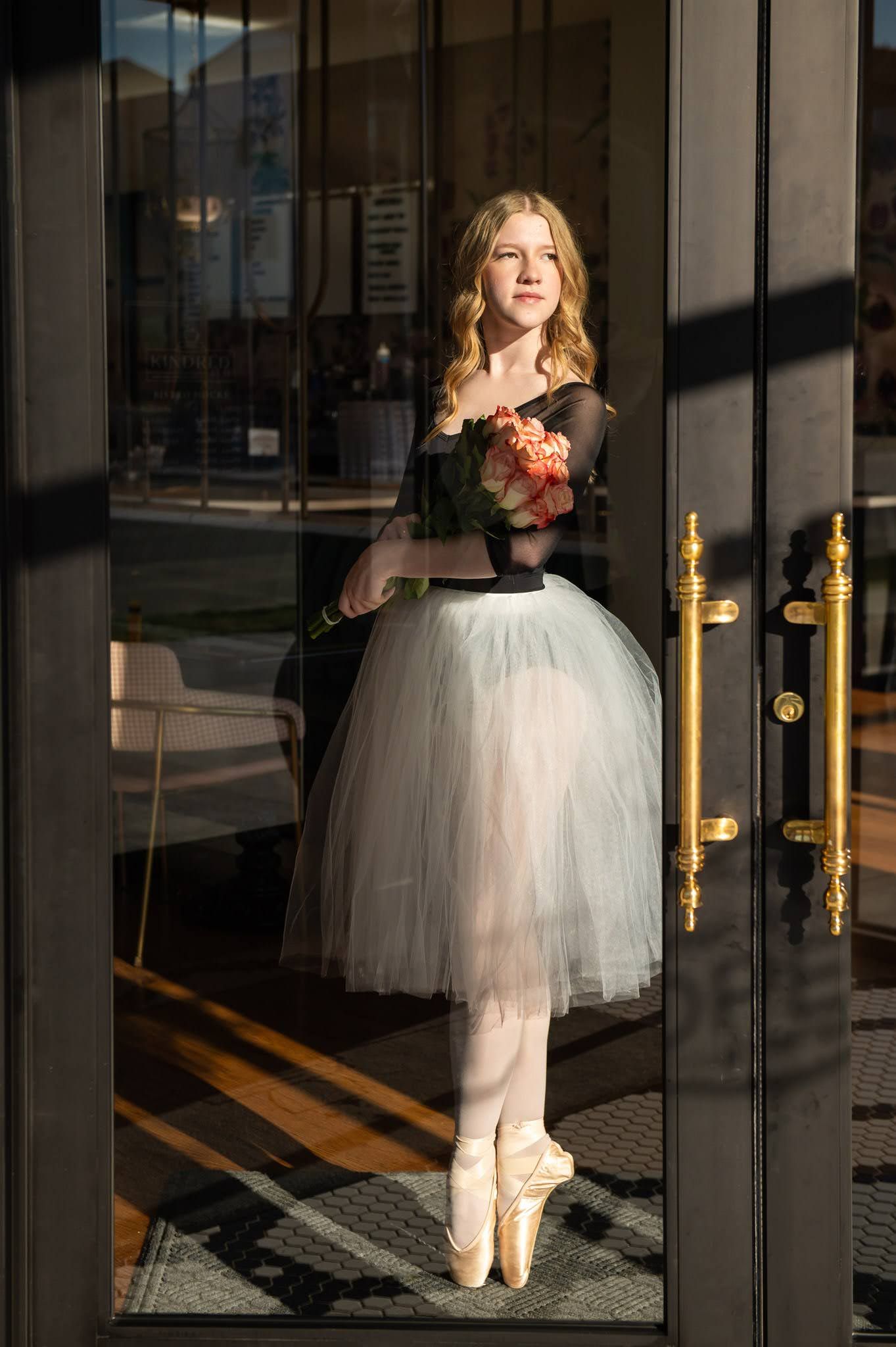 Elegant blonde ballerina in black top and gray tutu holding flowers standing en pointe in doorway.