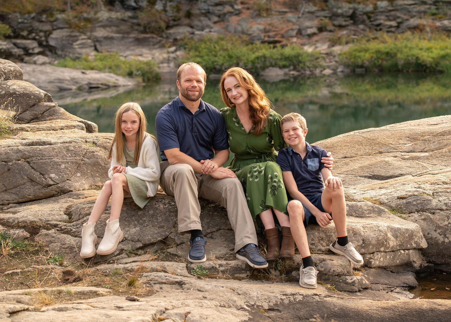 Family of four sitting on rocks near a scenic river outdoors.