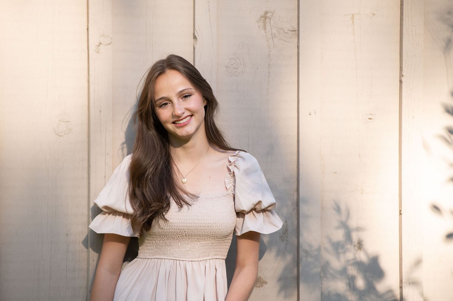 Young woman in a light pink smocked dress smiling against a wooden backdrop.