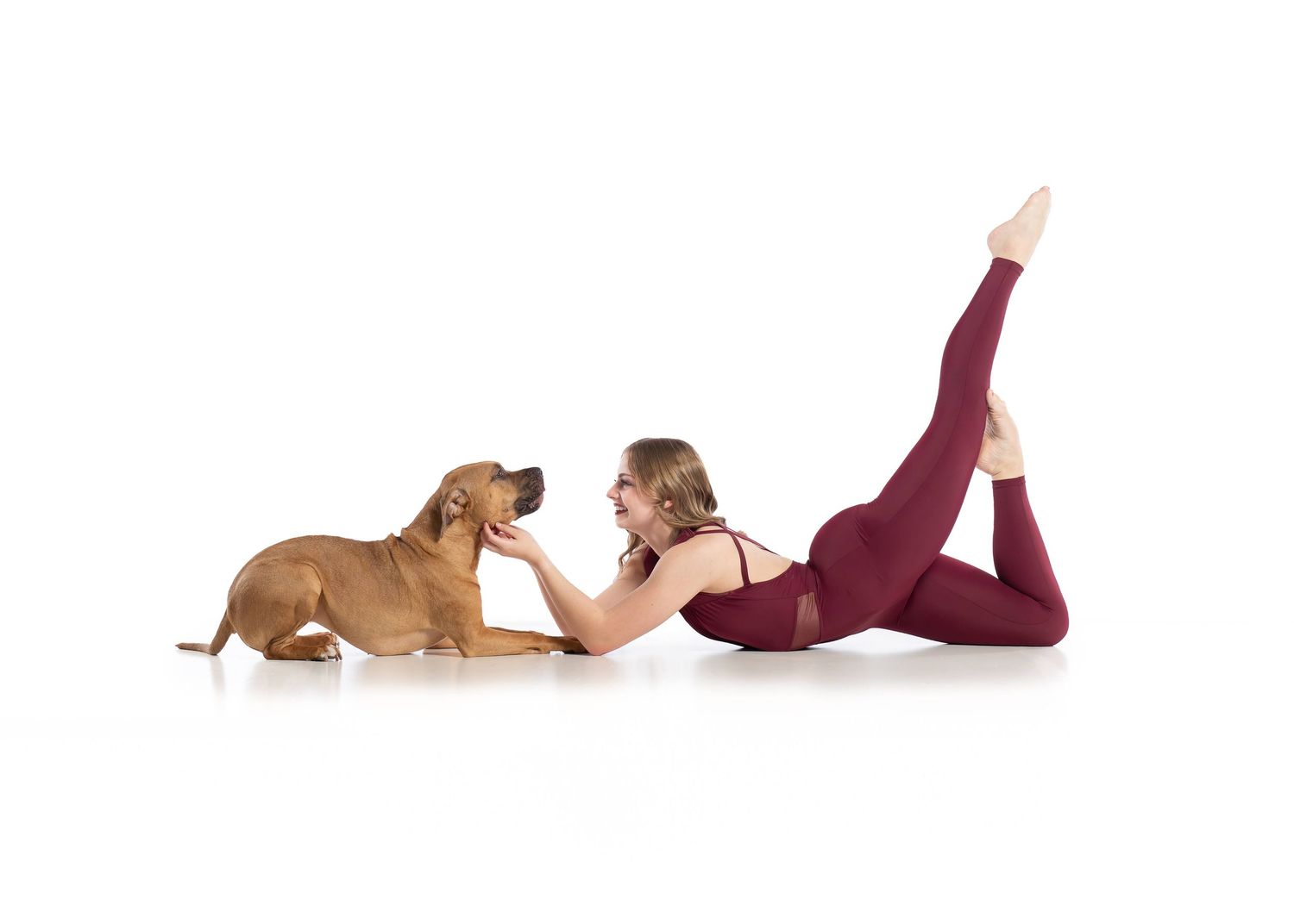 Woman in burgundy leotard lying on floor playfully interacts with a brown puppy on white background.