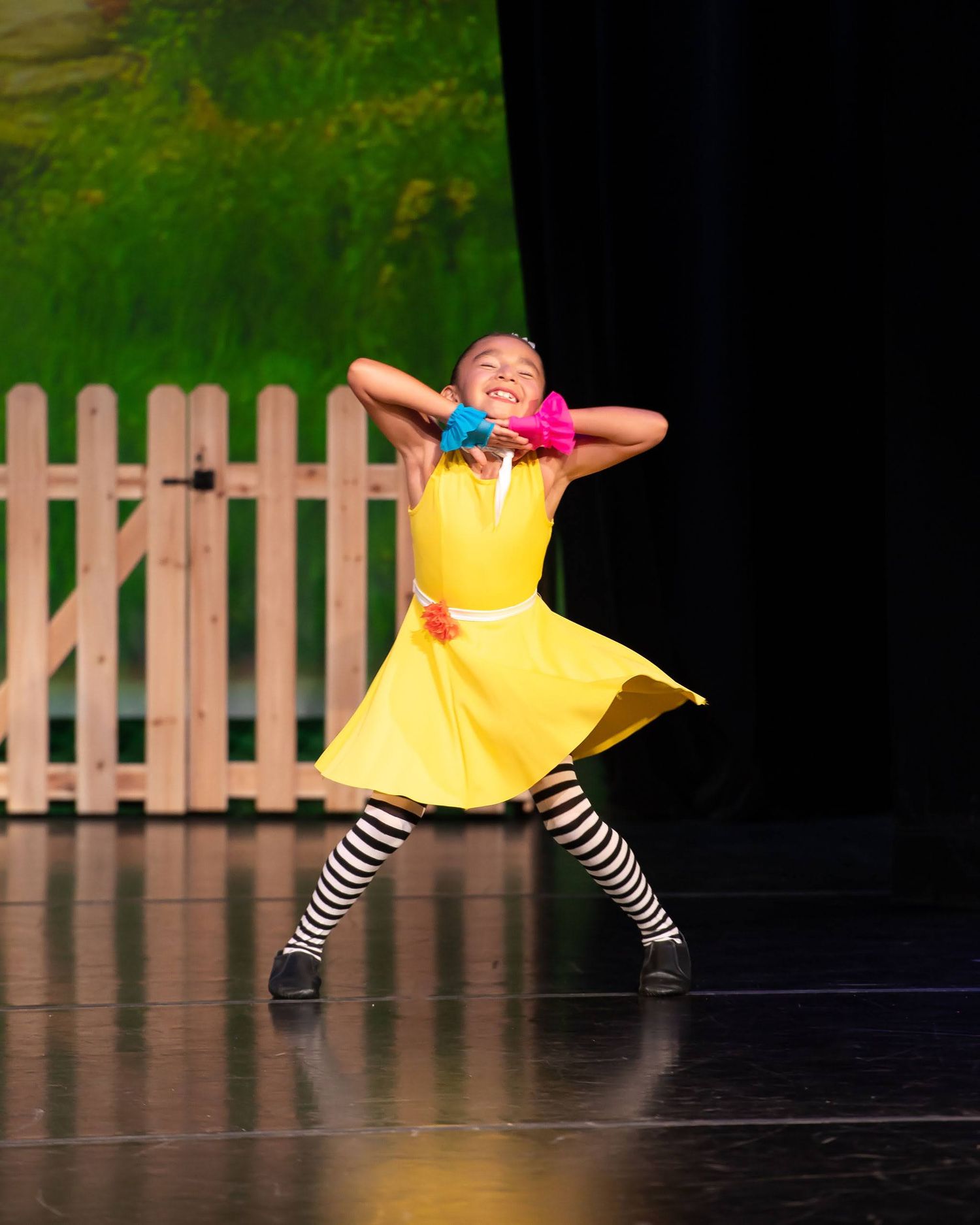 Young girl in yellow dress and striped socks performs energetic solo dance on stage.