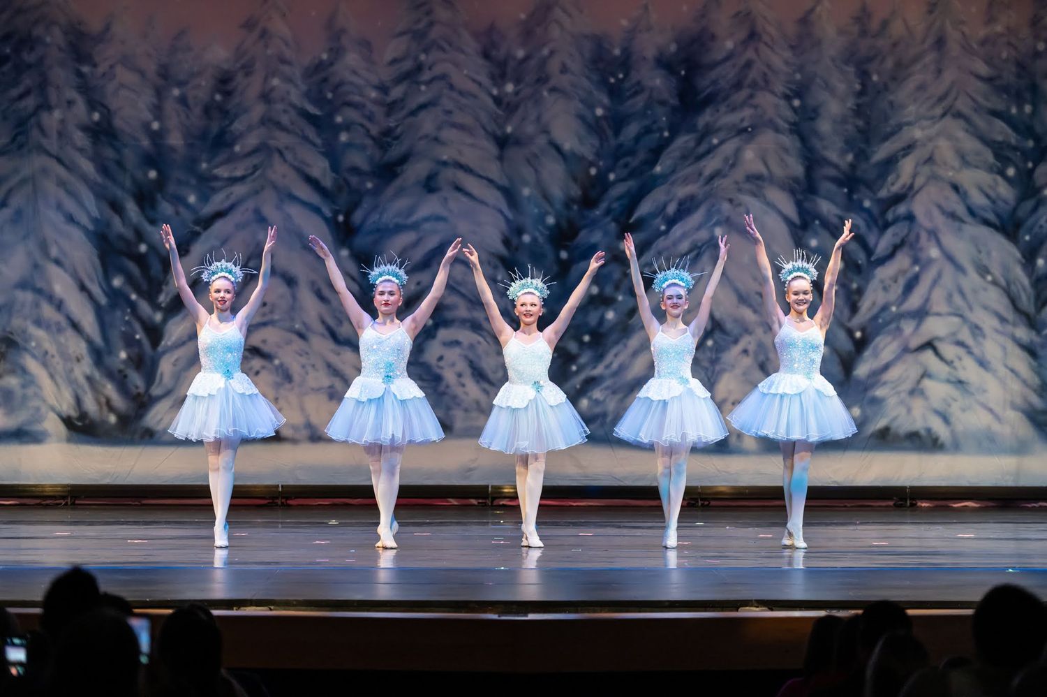 Five ballerinas in white tutus and blue crowns perform in unison before a snowy winter forest backdrop.