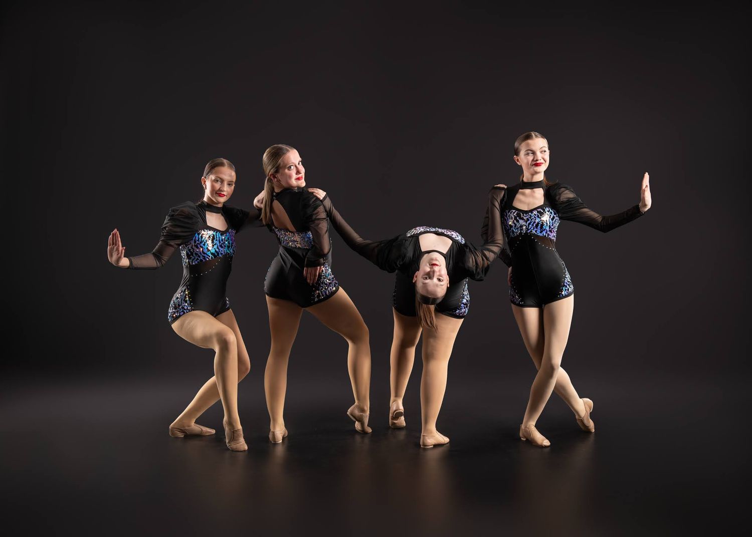 Four young dancers in black sequined leotards pose together in a dynamic group formation against a dark studio background.